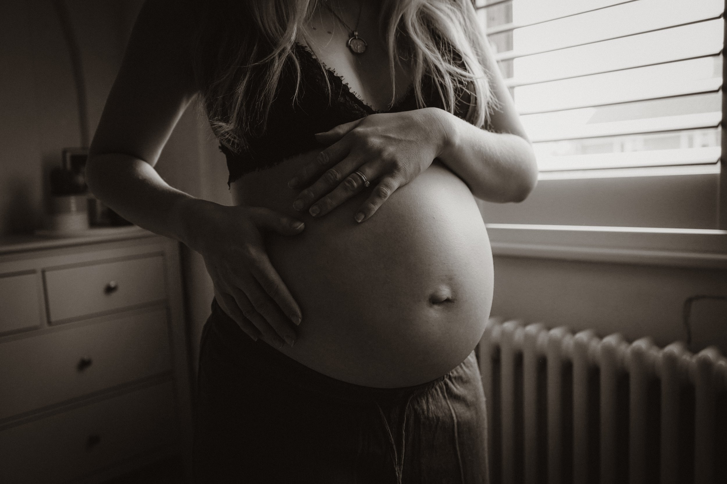 A black-and-white photo of a woman, focusing on her torso and hands, gently touching her pregnant belly near a window with blinds.