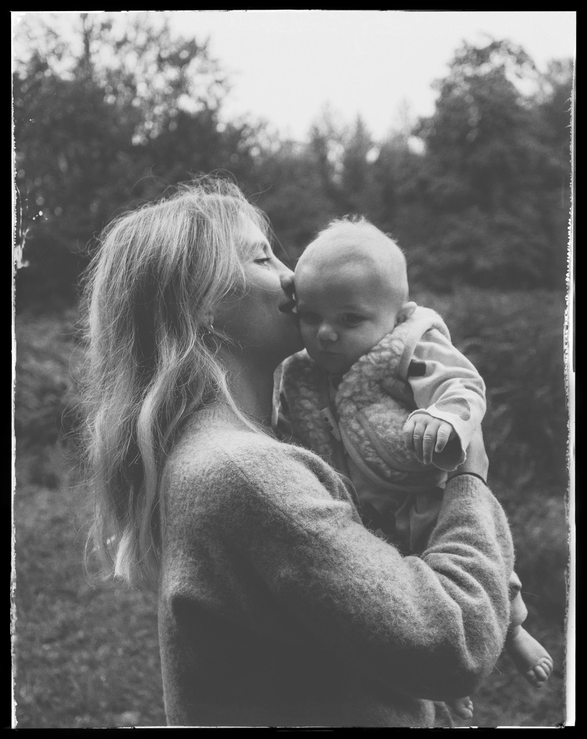 A woman with long blonde hair kissing a baby on the cheek outside in a park.
