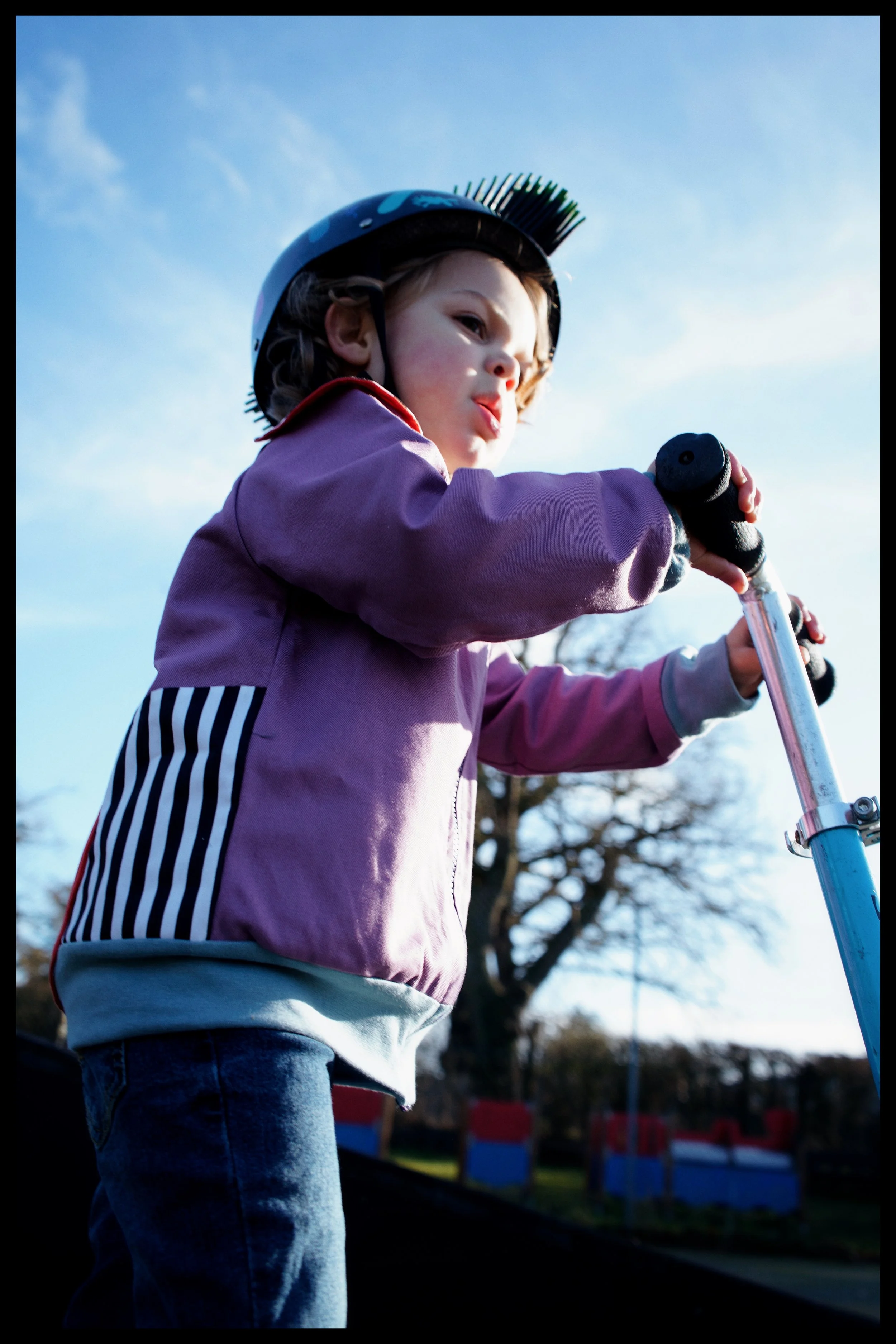 A young girl wearing a helmet with a brush attachment, riding a scooter outdoors on a clear day.