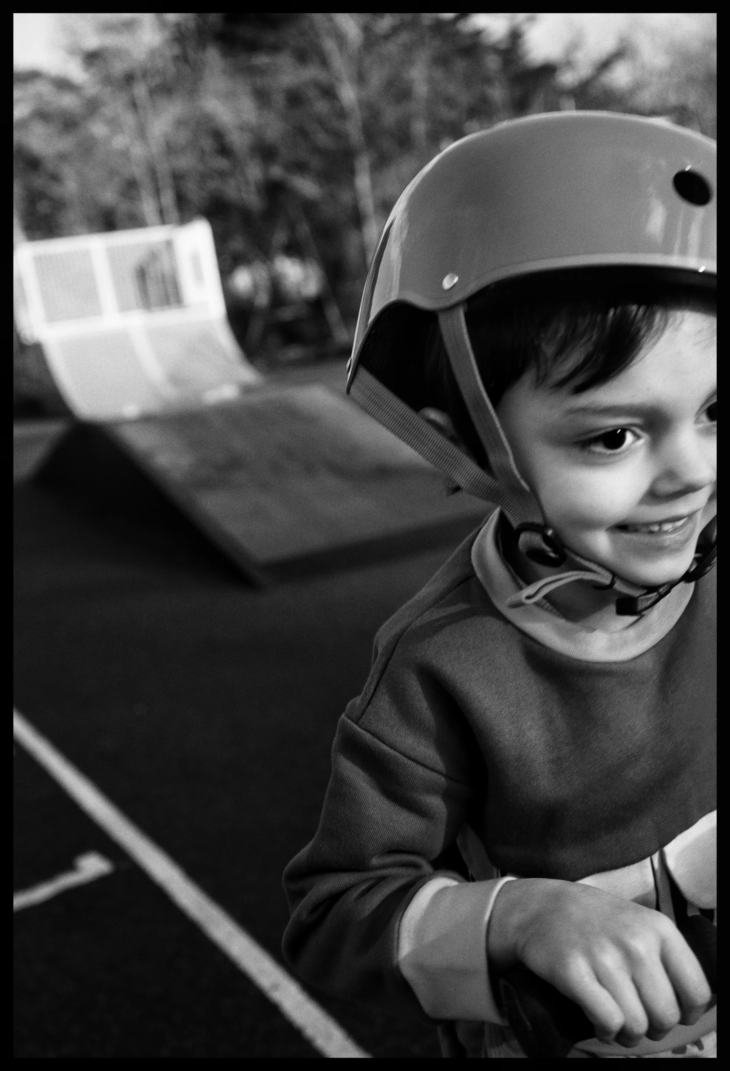 A young boy wearing a helmet and a jacket, smiling and sitting near a bowling lane with pins in the background.