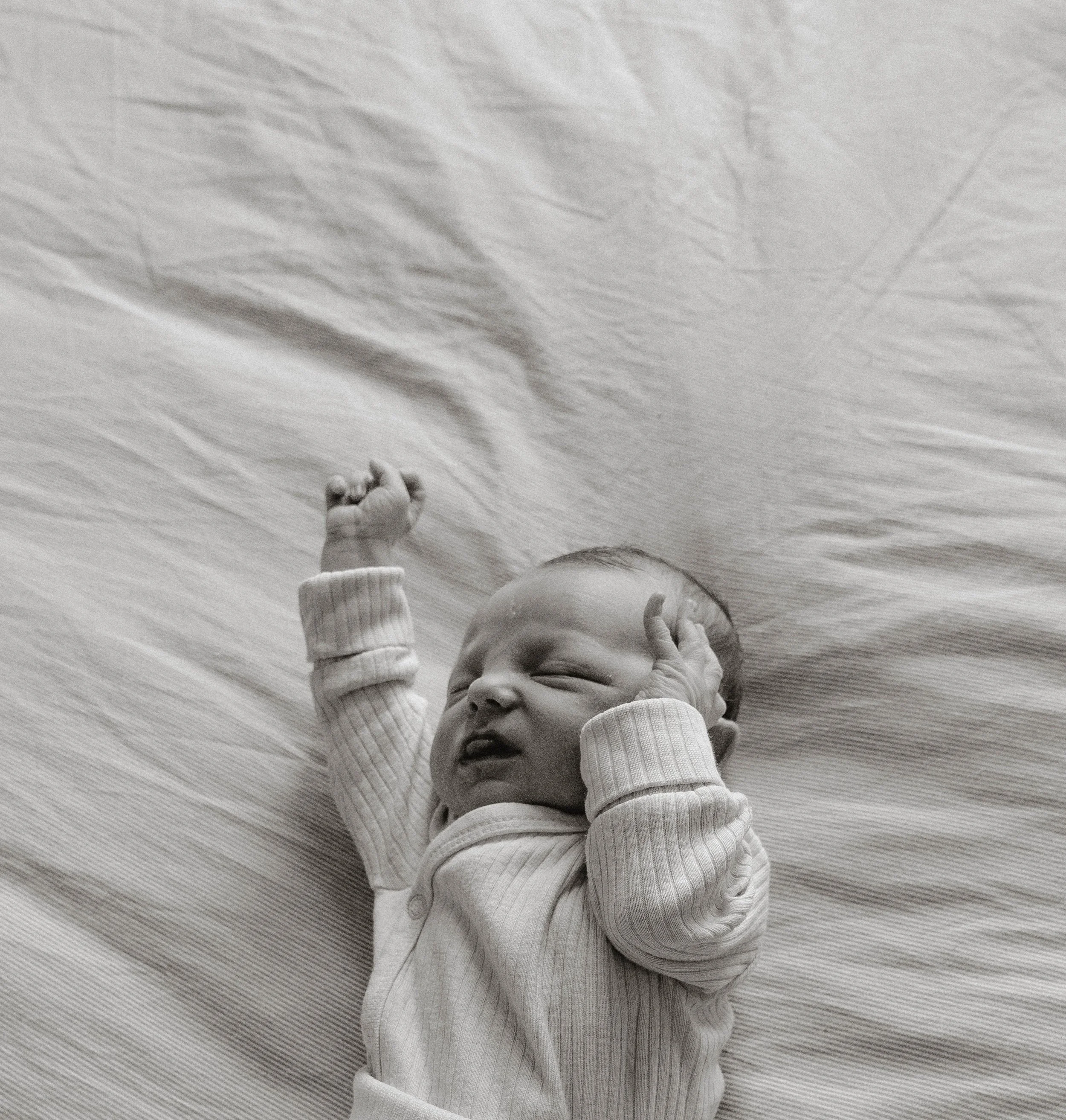 A baby lying on a wooden floor, wearing a striped long sleeve shirt and pants, with one hand raised and the other near their face, making a face with their mouth open.