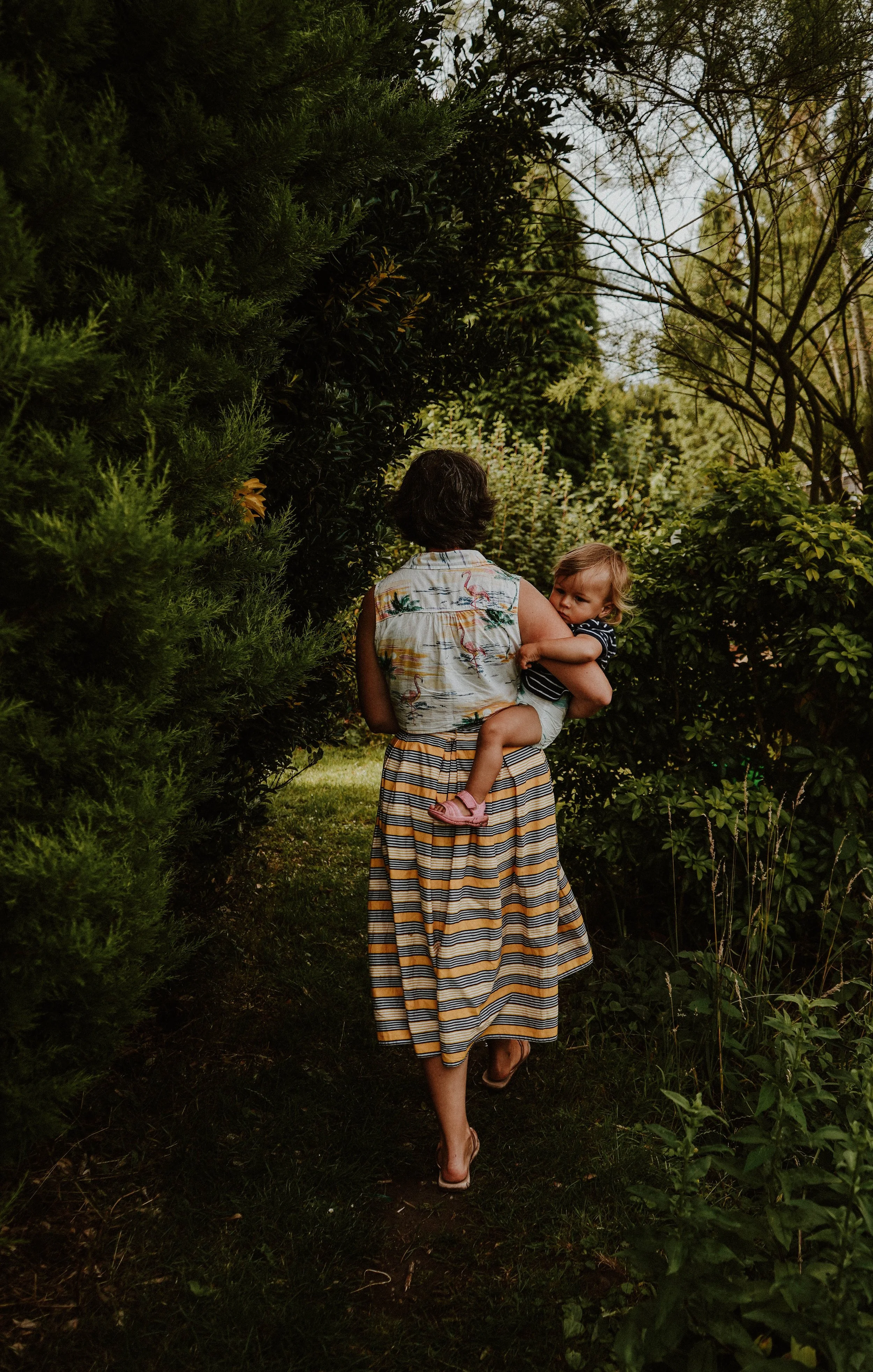 A woman walking through a lush garden or park holding a young child. The woman is wearing a striped skirt and a sleeveless tropical shirt. The child is in a striped shirt and shorts, resting in the woman's arm with a curious expression. Surrounding t