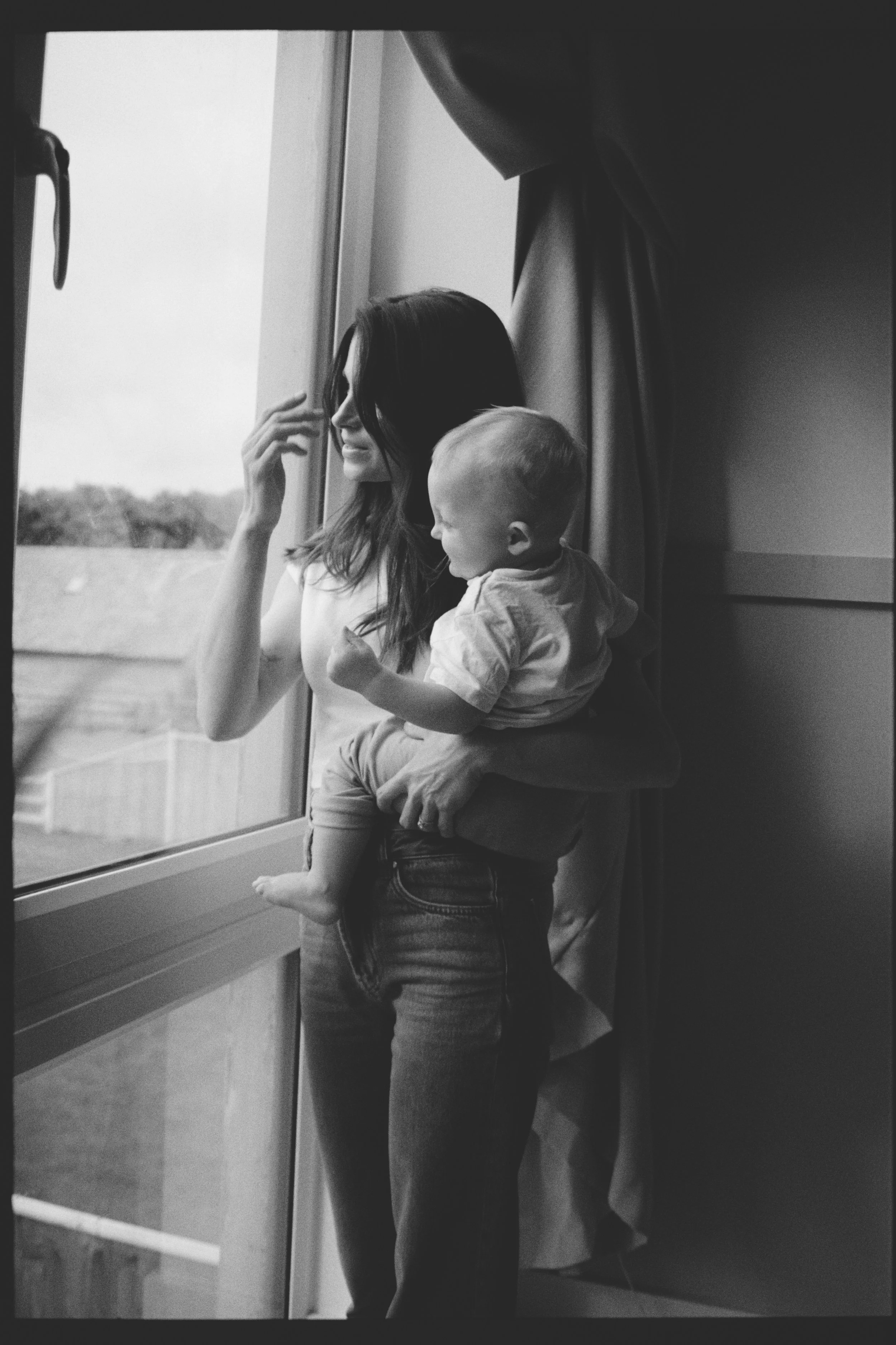 A woman with long dark hair holding a young boy with light hair, both looking out of a large window. The woman is touching her forehead with her right hand, and the boy is sitting on her waist with his legs hanging down. The scene is in black and whi