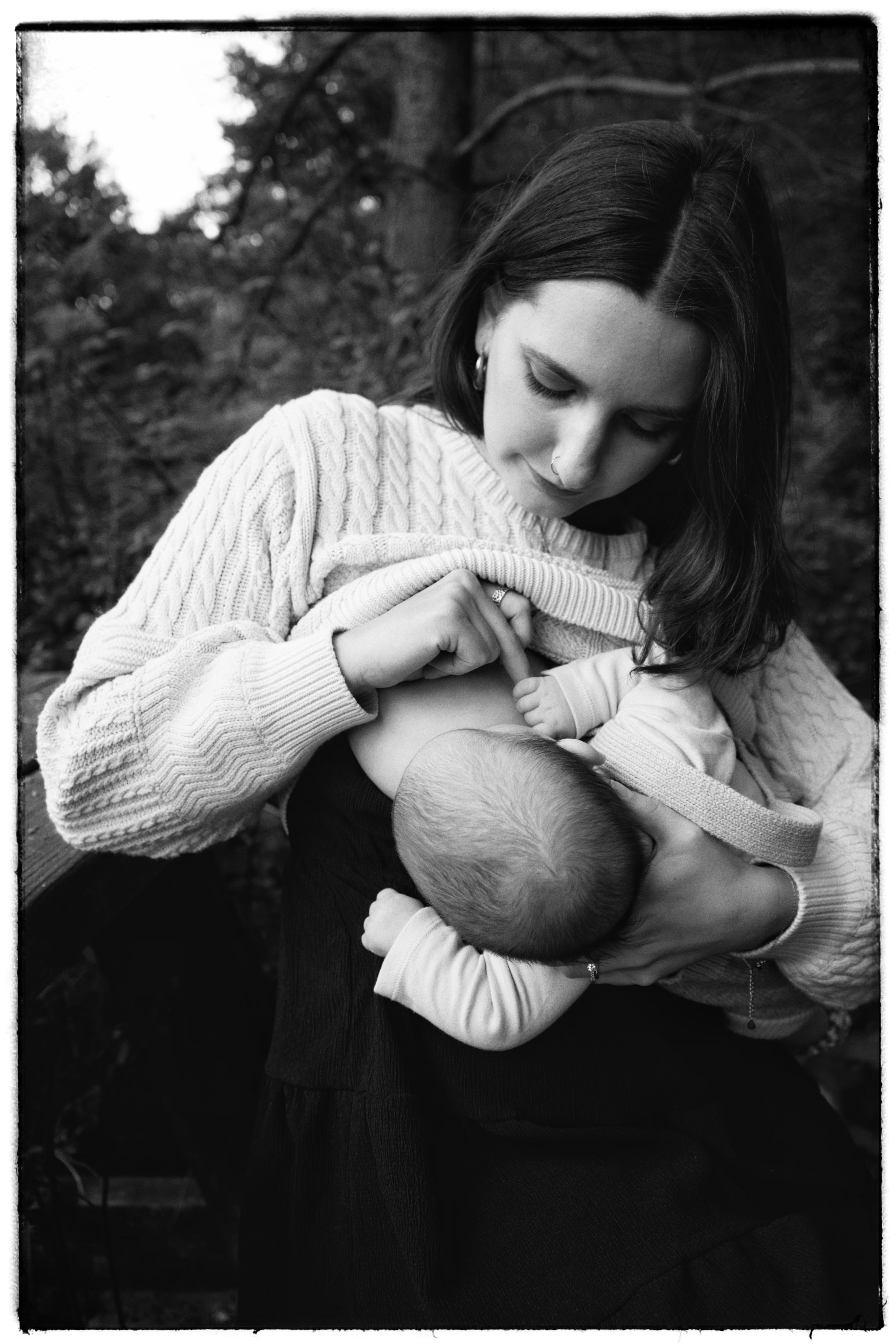 A woman with dark hair, wearing a sweater, holding a baby in her arms, outdoors with trees in the background, in black and white.