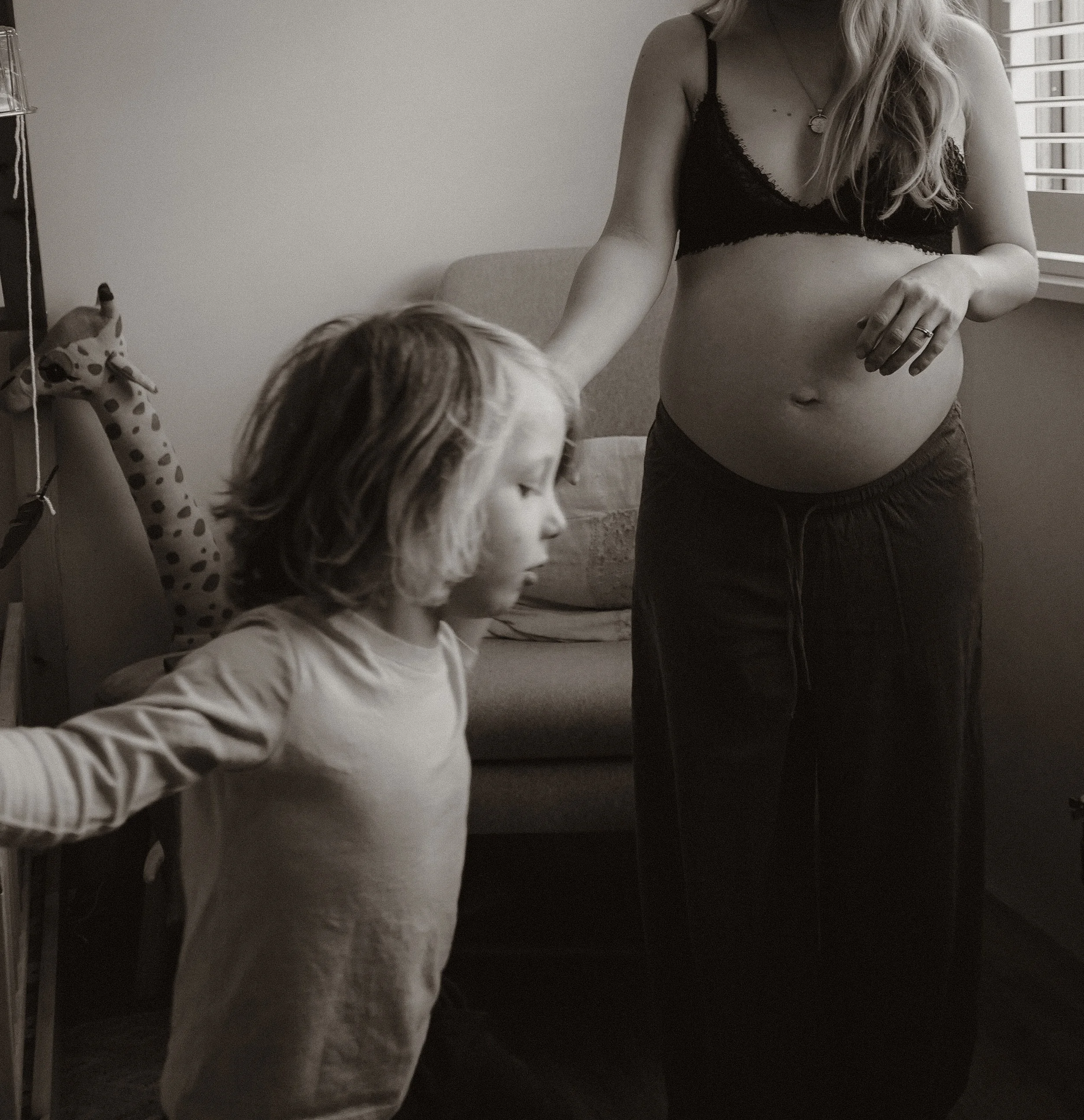 Girl reaching for giraffe plush toy on a shelf in a room with a window and a person with long hair and a pregnant belly.
