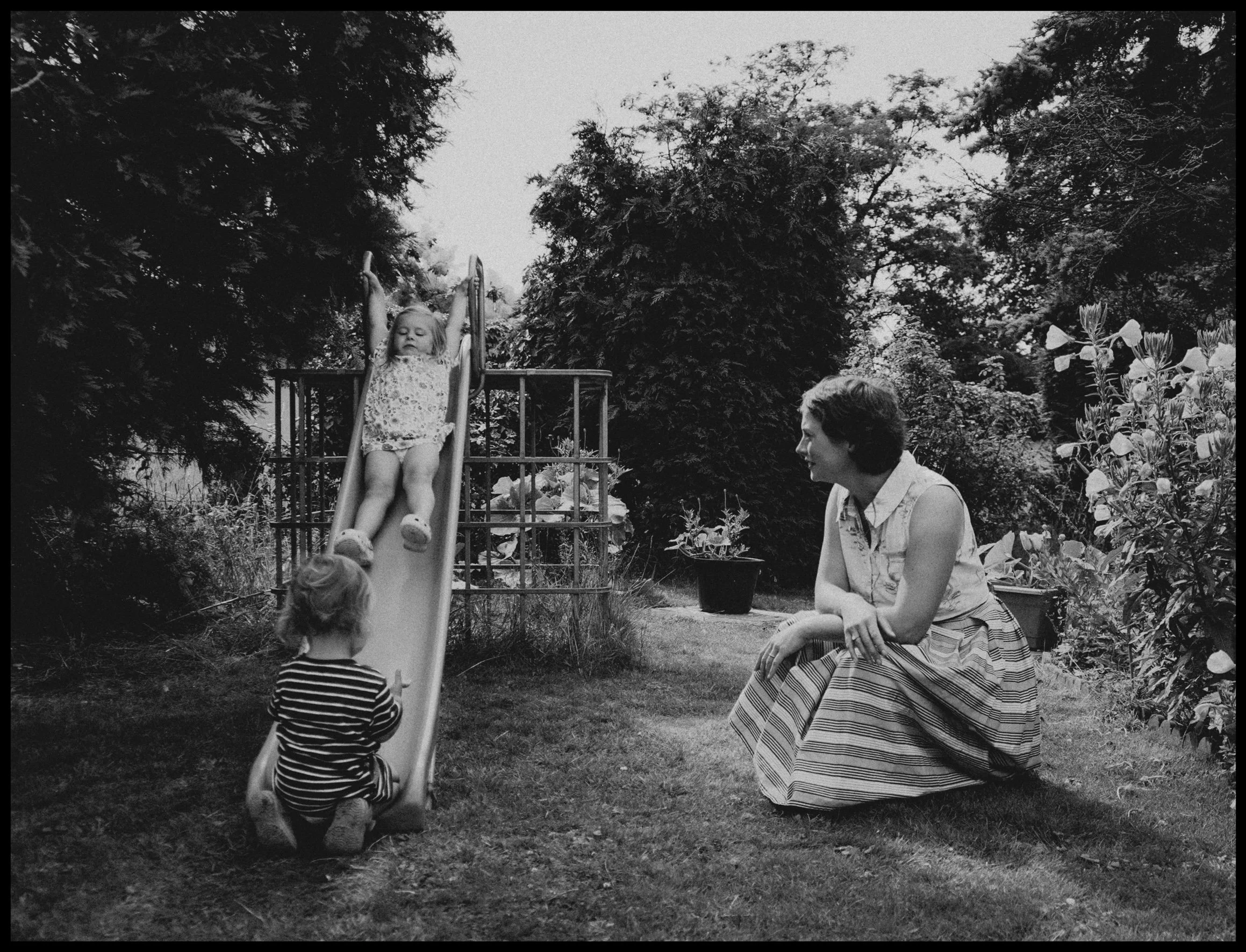 A black-and-white photograph depicting children and women in a garden or backyard setting. A girl is coming down a slide, and two other children are nearby on the grass. One woman is sitting on the grass, while another woman stands, watching the chil
