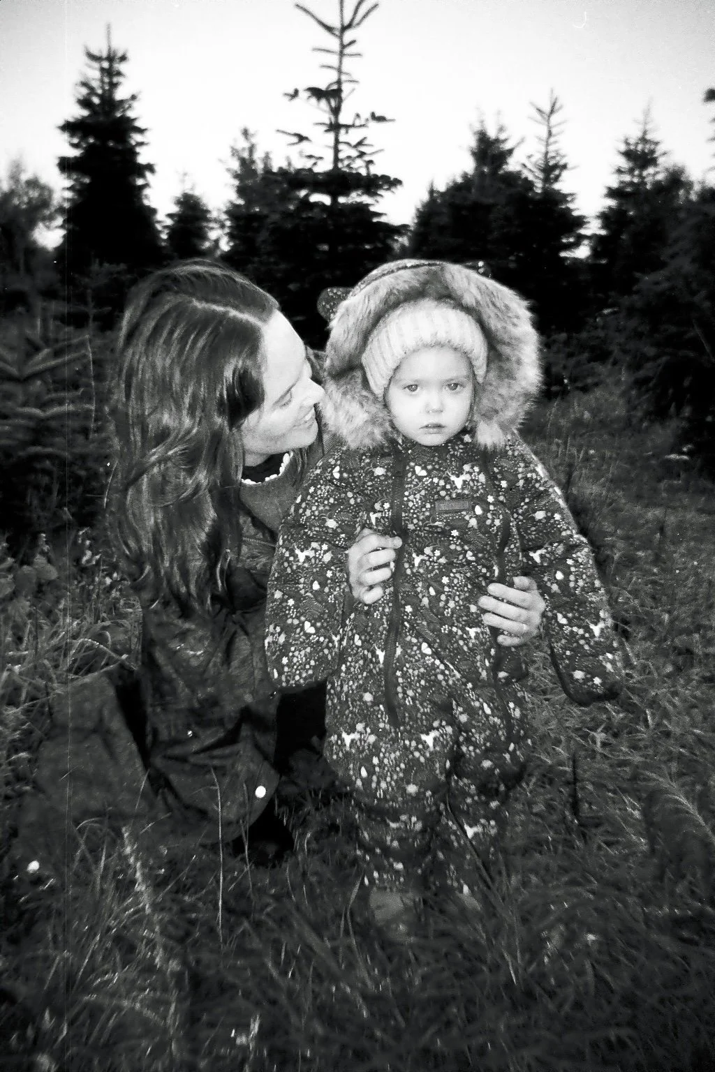 A woman crouching next to a young girl in a forest, both wearing winter jackets and the girl in a fur-lined hood and knit hat, captured in black and white.
