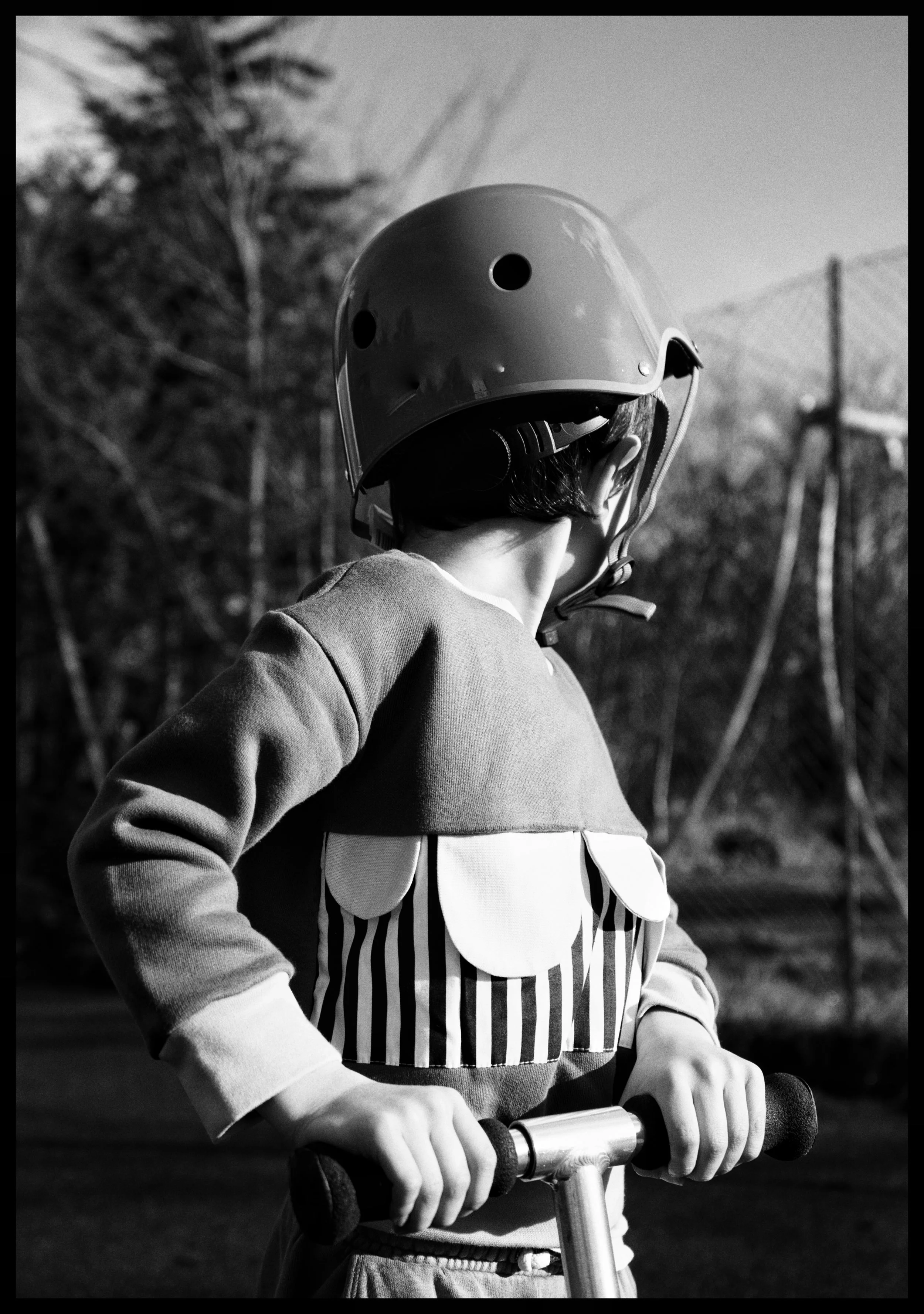 Child wearing a helmet, standing outdoors, holding the handlebars of a scooter, with trees and a fence in the background.