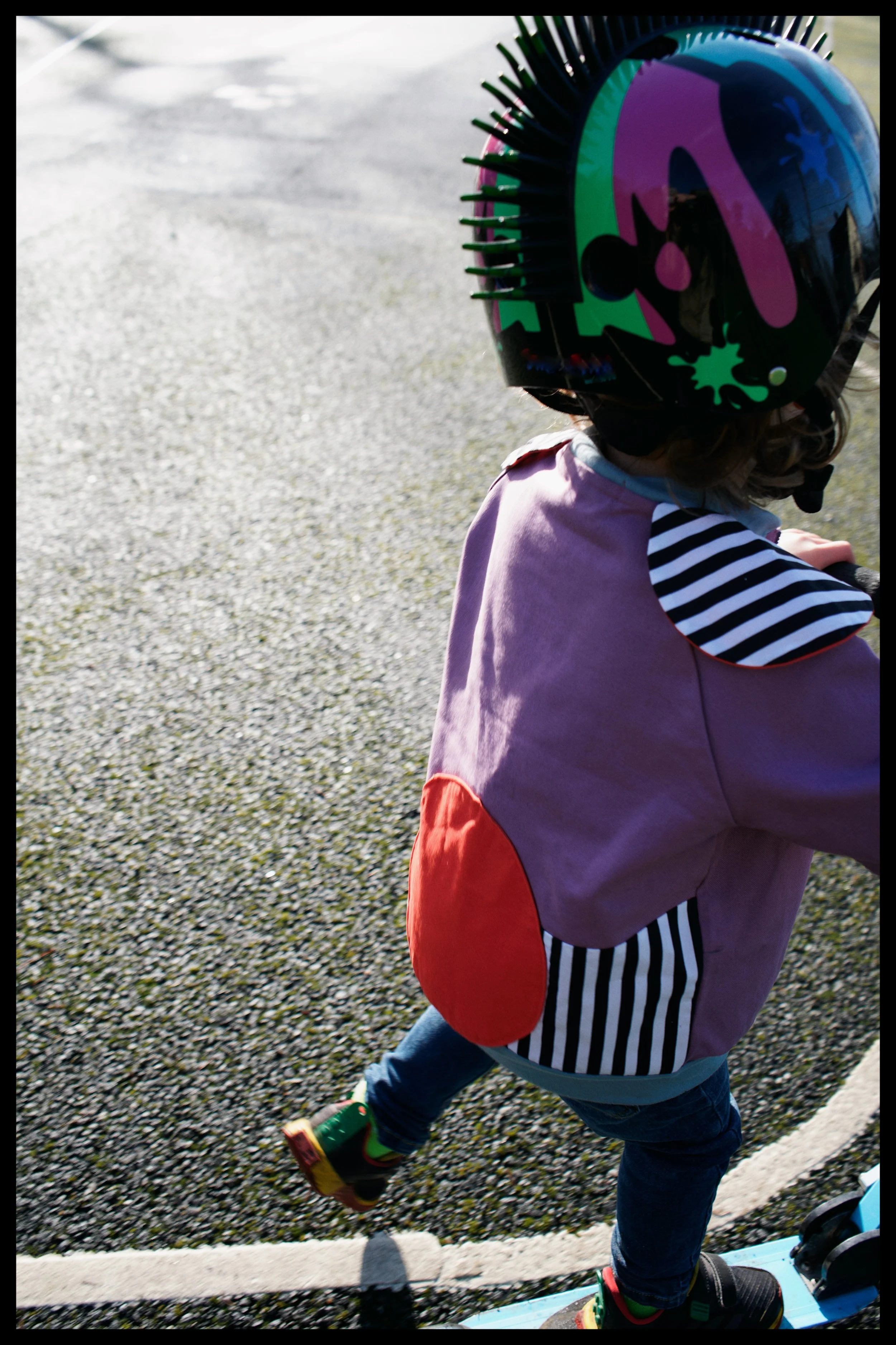 A child wearing a helmet and colorful jacket riding a skateboard outdoors on a paved path.