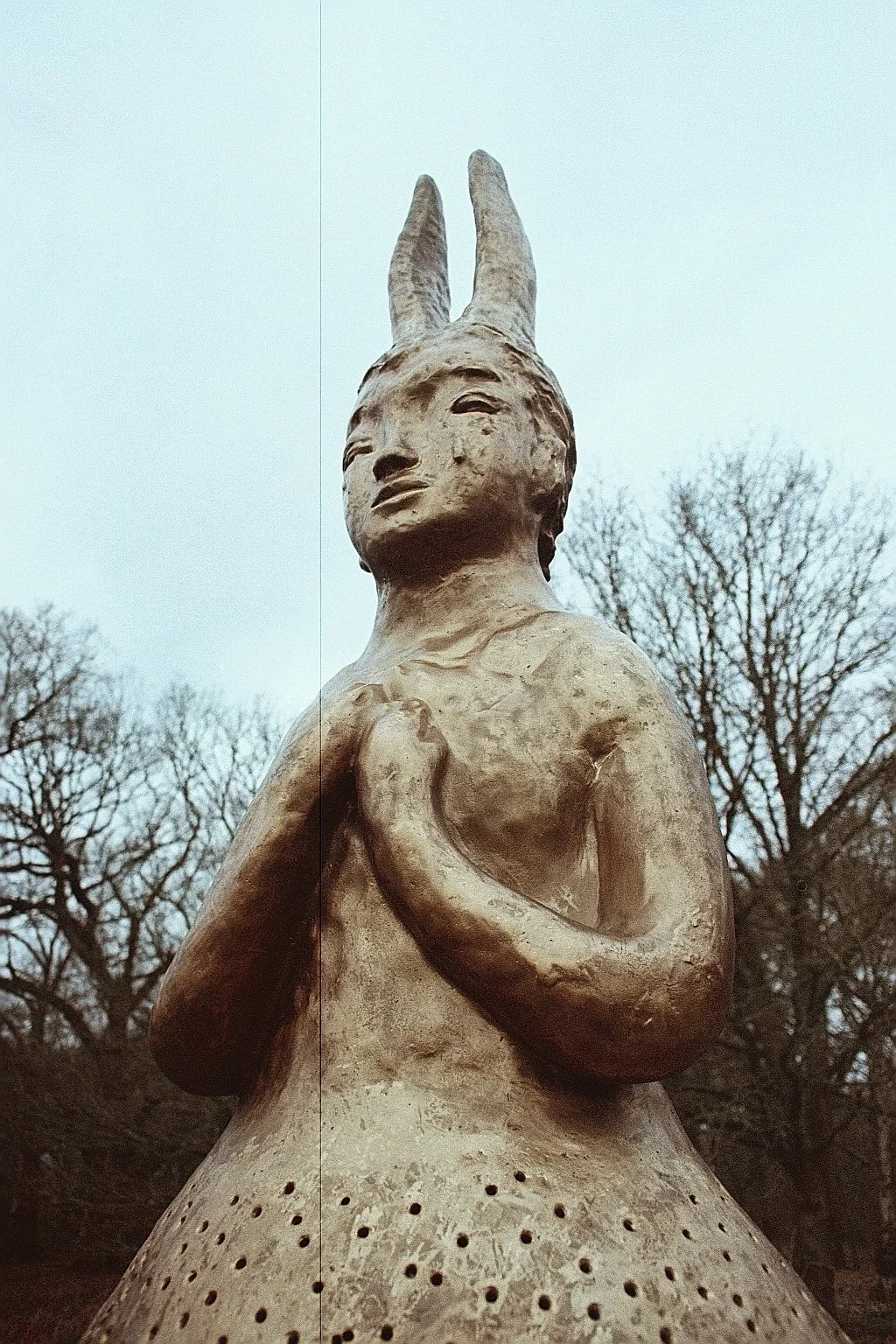 Scenic outdoor statue of a woman with a halo and horns, holding her hand to her chest, with leafless trees in the background.