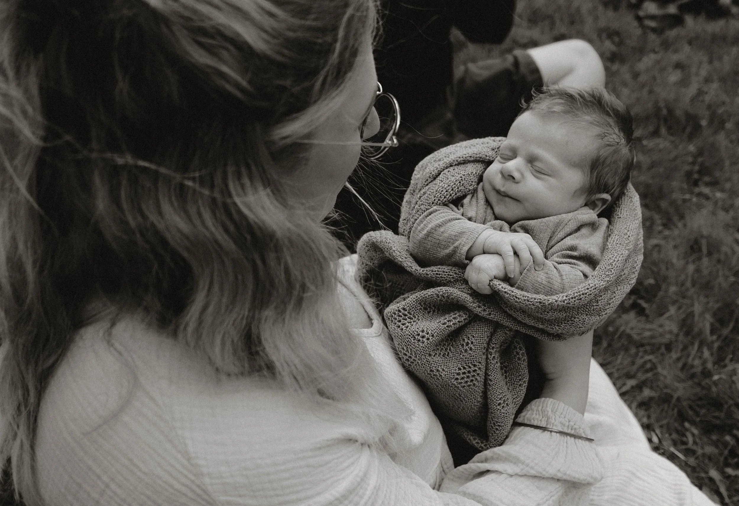 A woman with long hair and glasses holding a smiling baby in her arms outdoors.