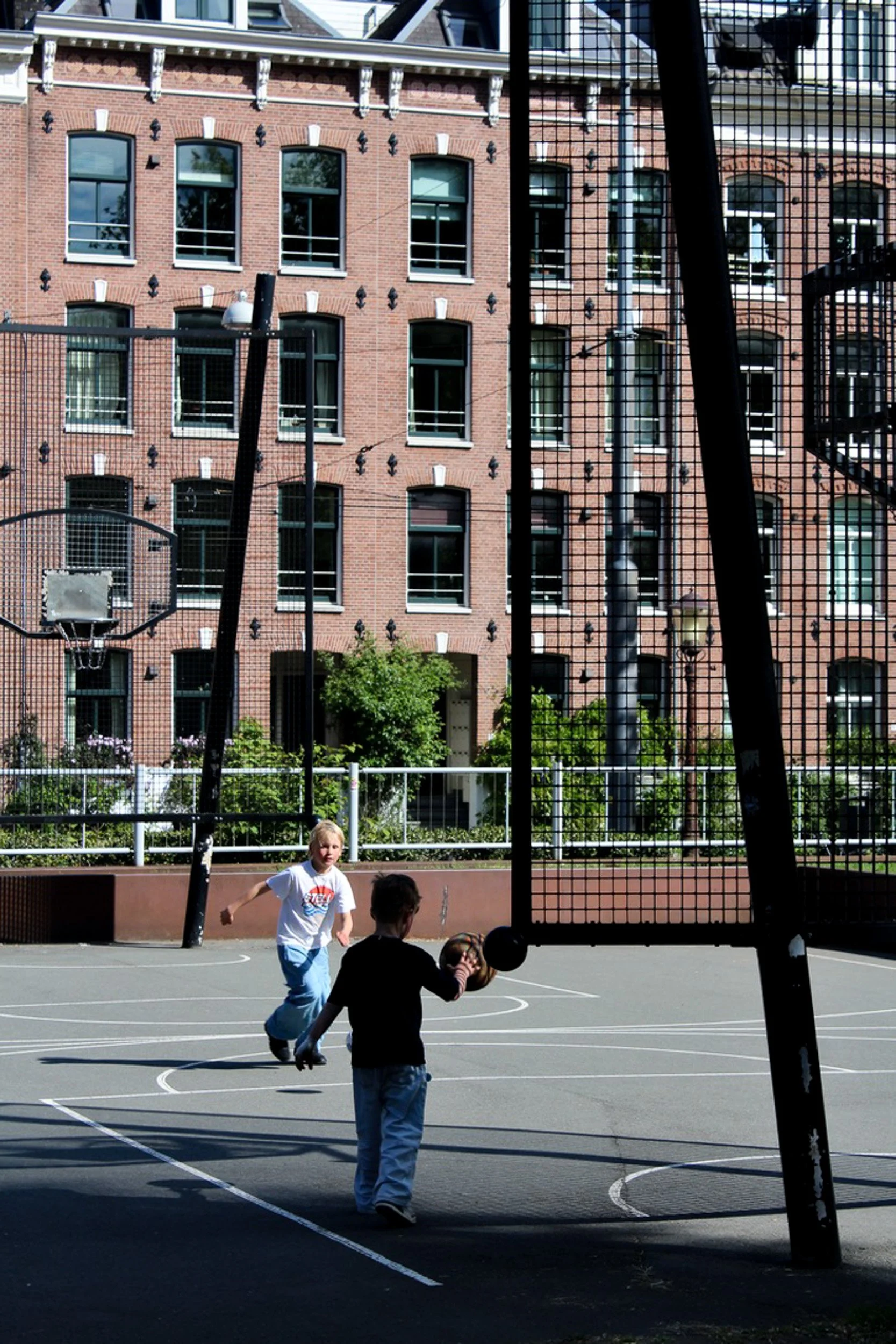 Children playing basketball on an outdoor court in Amsterdam, photographed through a fence. Street photography by Isabel Hachmann-Peralt.