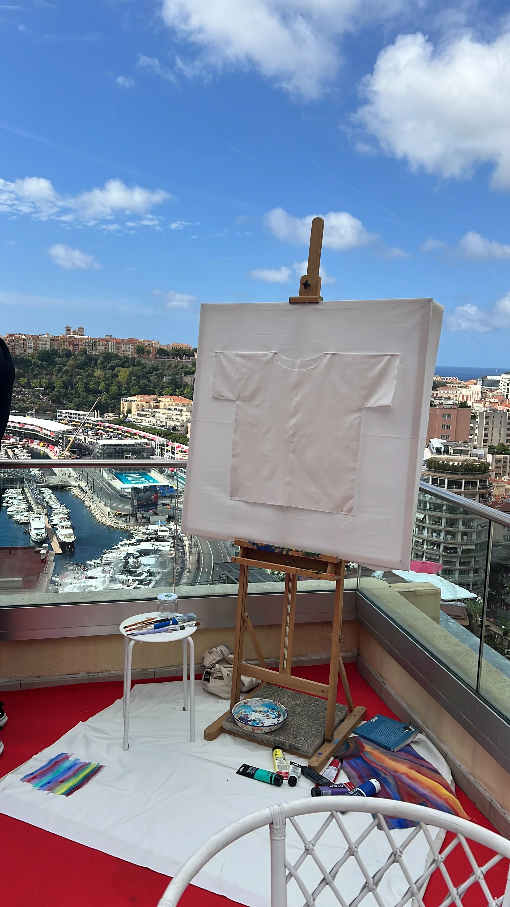 An artist's easel on a balcony with a city and marina view behind, painting supplies on a small white table, painting materials and a canvas on the floor, with blue sky and some clouds overhead.