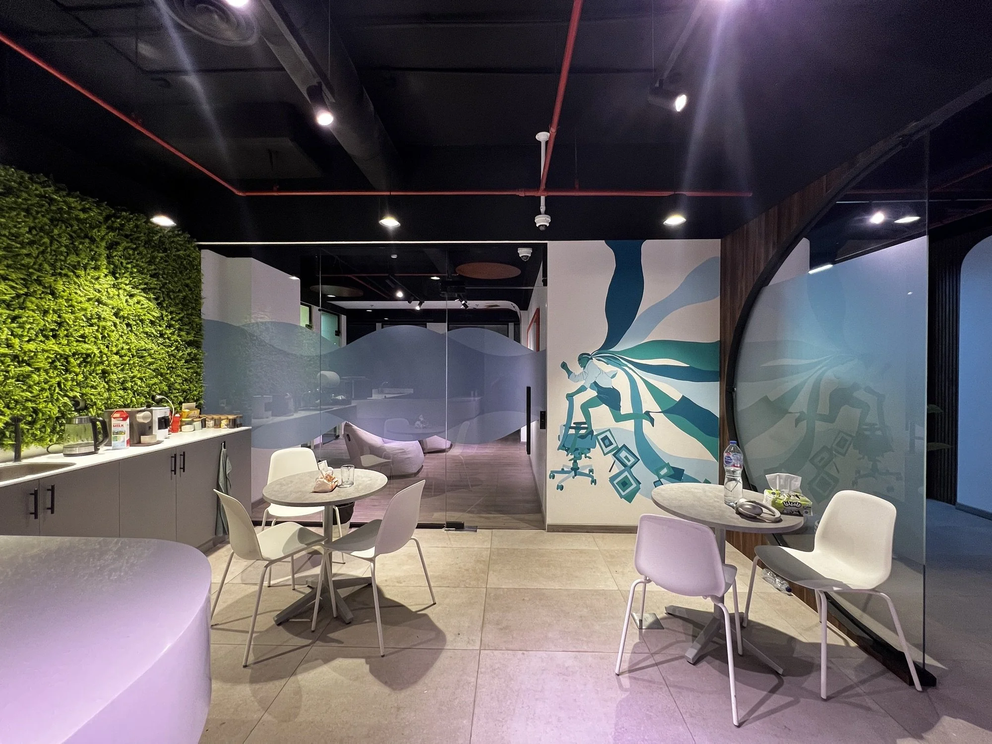 Interior view of a modern office or break area with round tables and white chairs, a wall mural of a dancer, green wall plants, and a beverage station with coffee supplies.