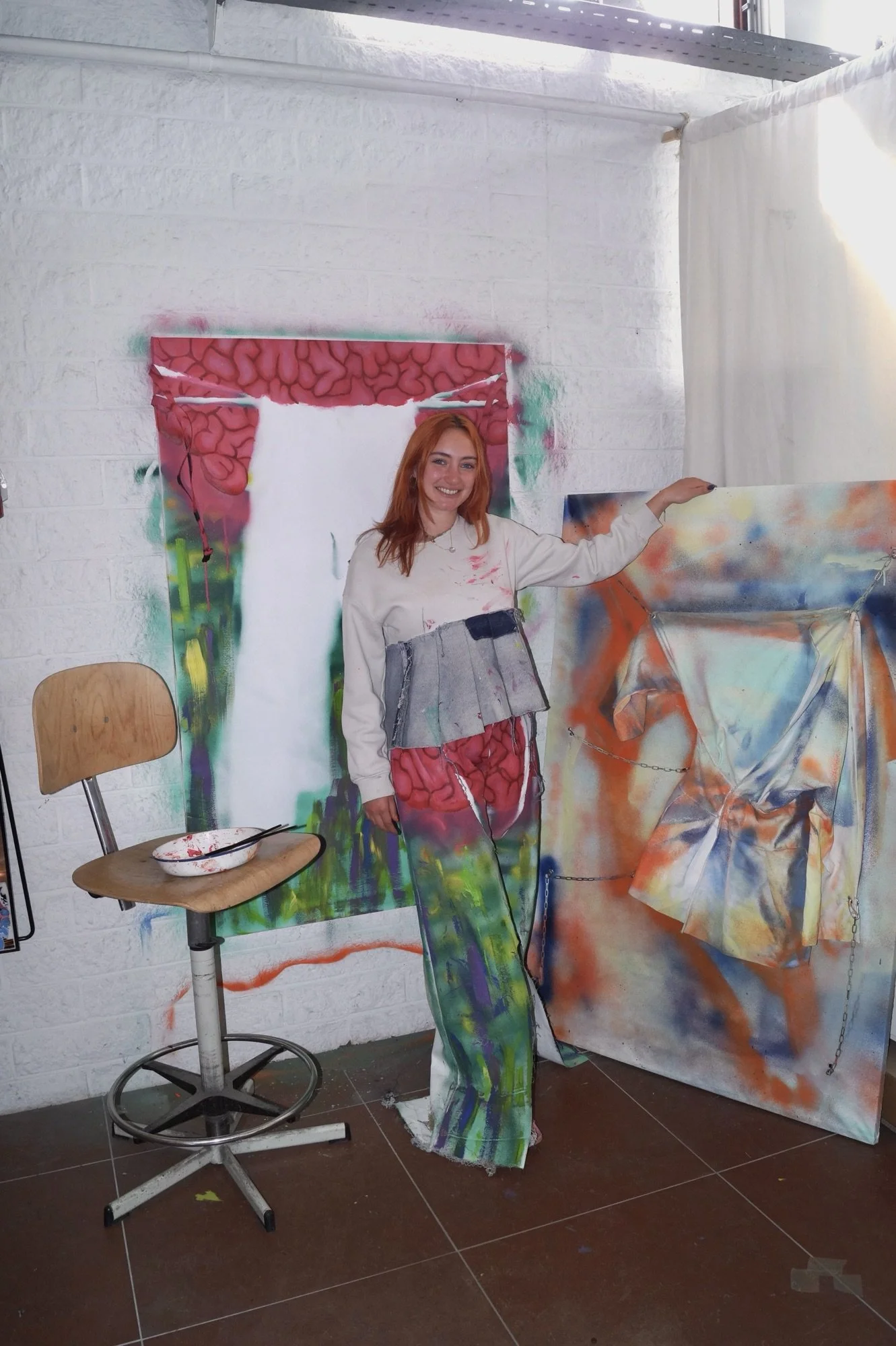 A woman with orange hair standing inside a colorful art studio, wearing a dress that looks like a piece of art. She is smiling and standing next to a large, abstract painted canvas, with a background of street art on the wall.