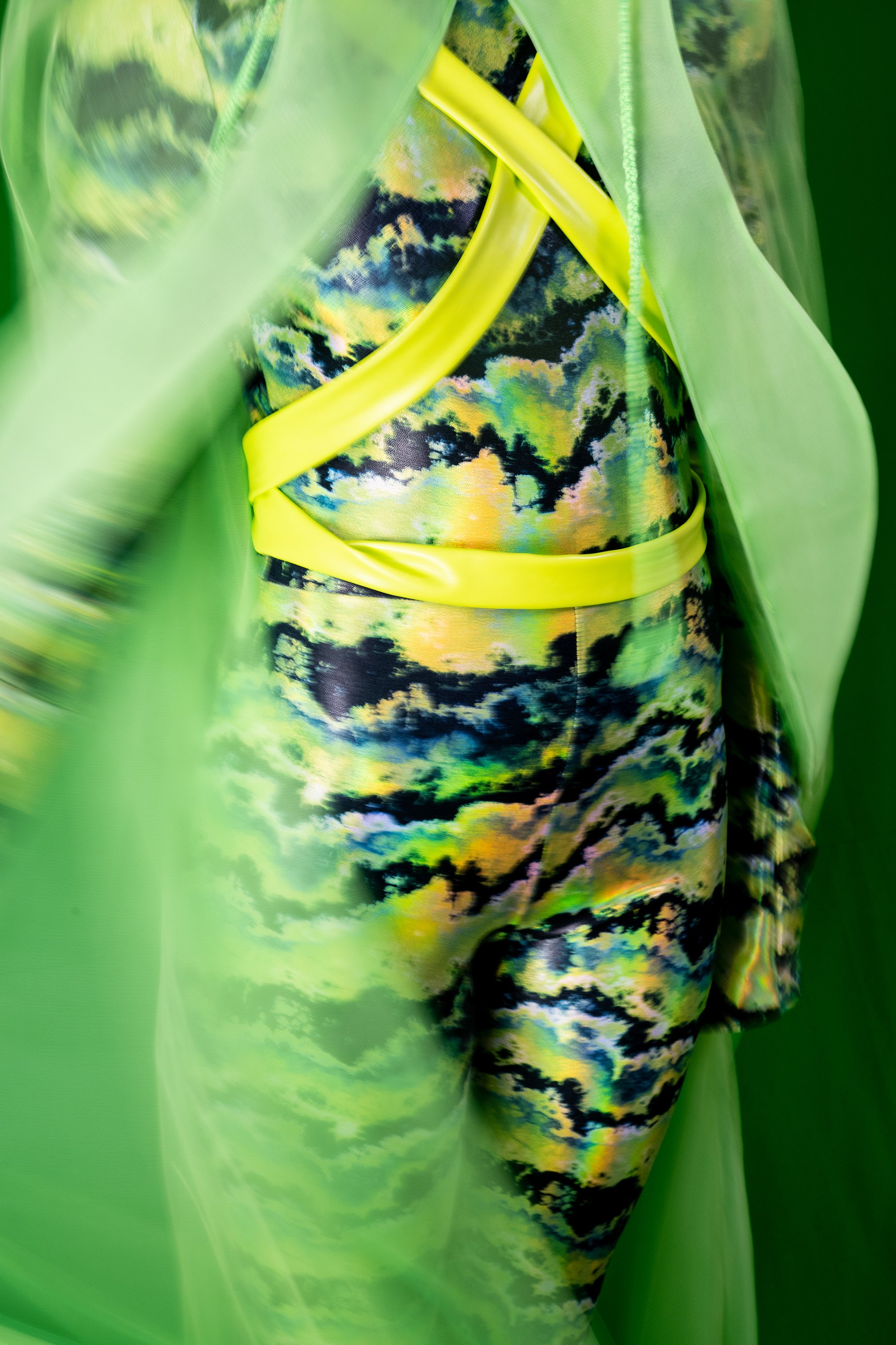 A person wearing a dress with a vibrant, multicolored cloud pattern, wrapped with neon yellow straps, surrounded by green translucent fabric.