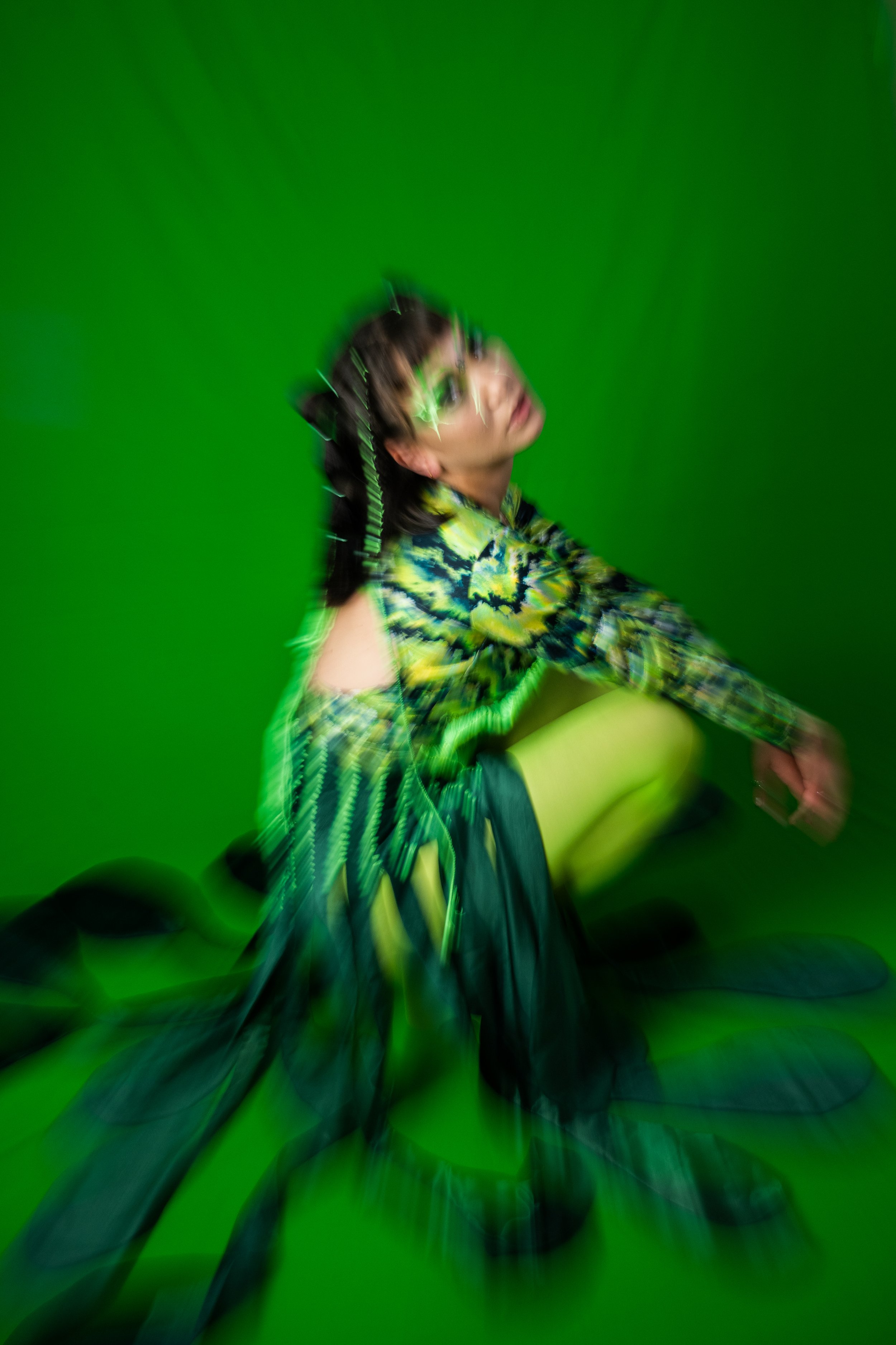 A person sitting against a green screen background, wearing a colorful, patterned shirt with a plaid design in green, blue, and black, and neon green tights. The photo has a motion blur effect.