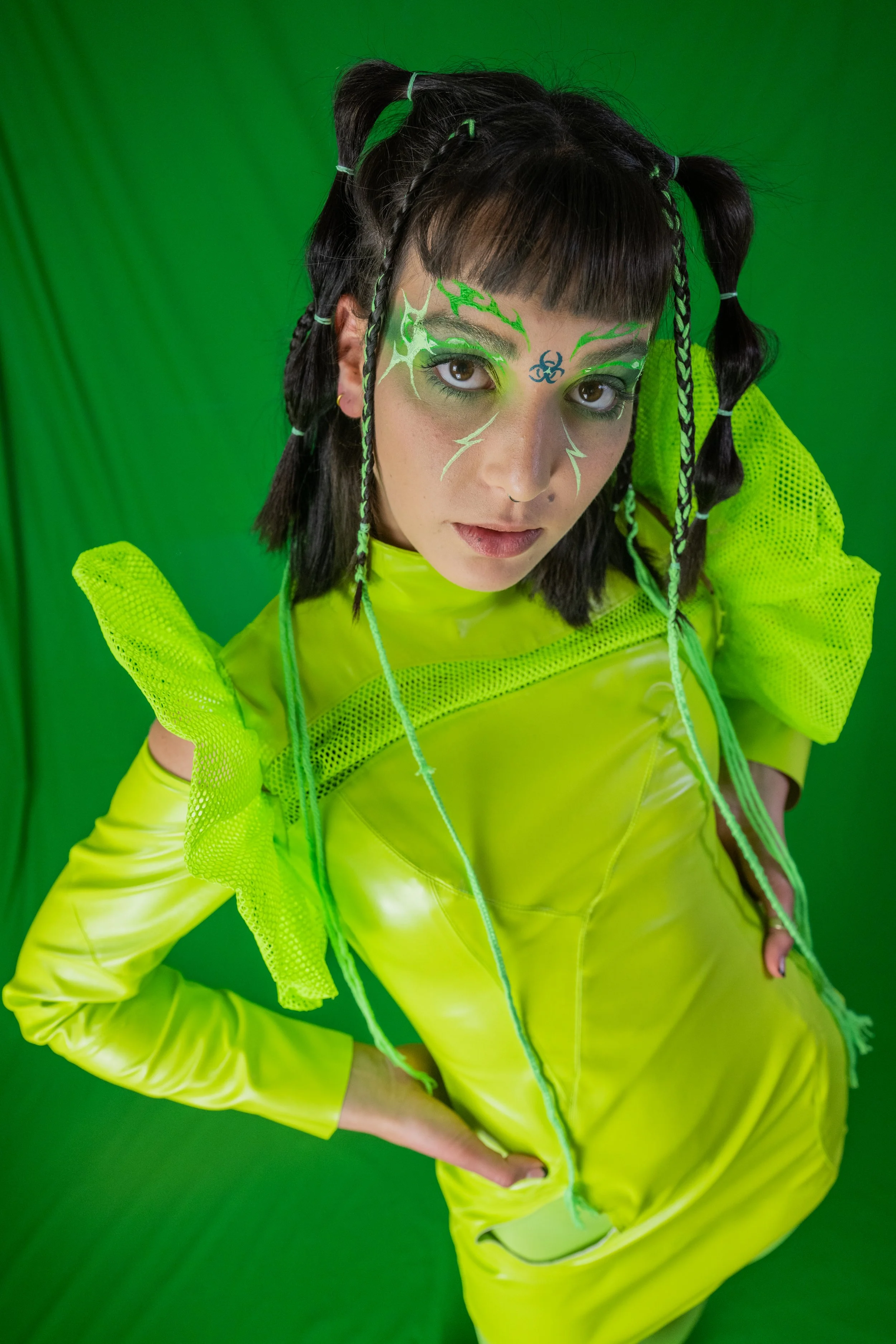 A young woman with dark hair styled in braids and pigtails, adorned with neon green hair ties. She has facial makeup with green and blue biohazard-themed designs, including a biohazard symbol on her forehead. She is dressed in a bright yellow futuris