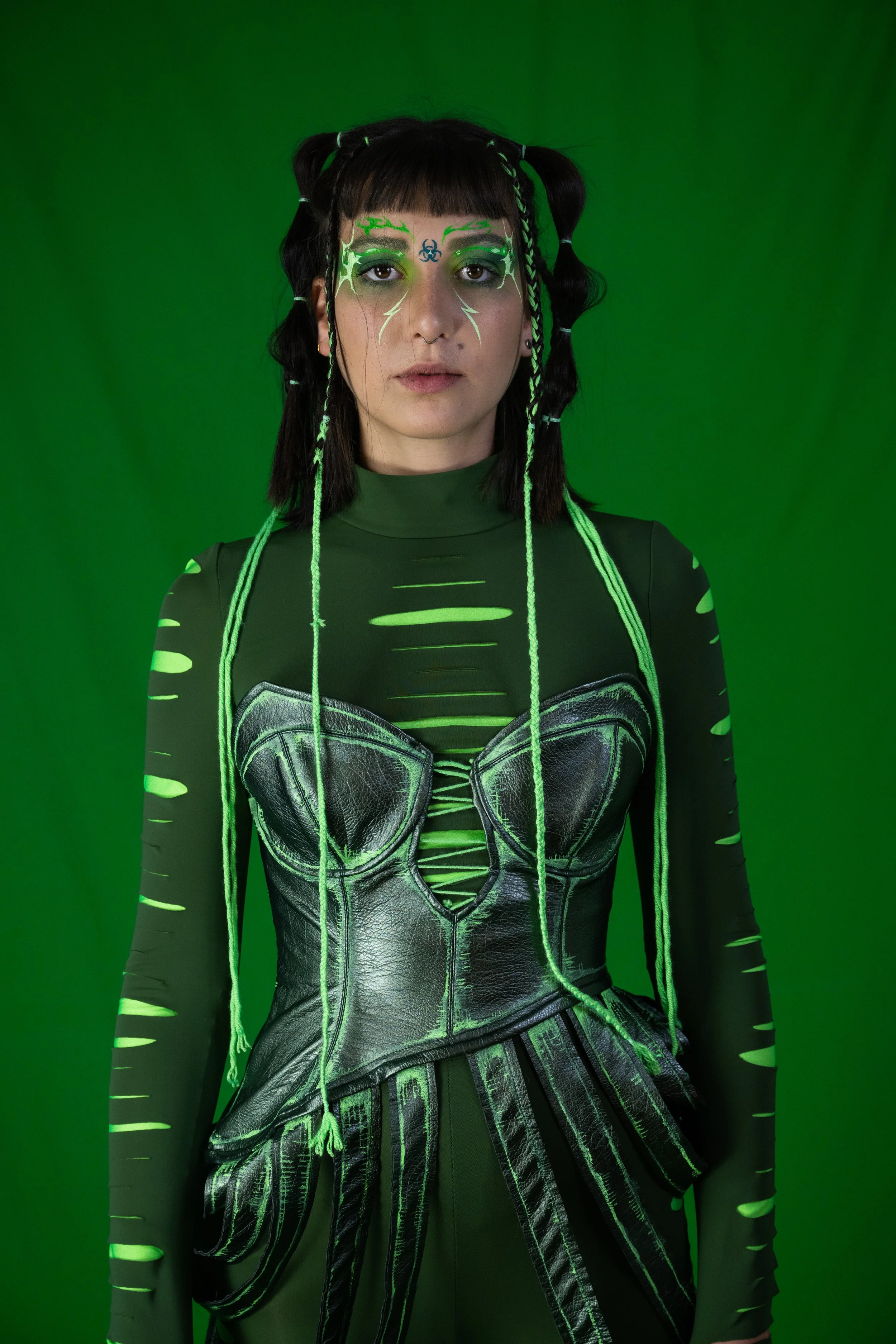 Young woman with black hair styled in multiple pigtails with green and white hair ties. She wears neon green face paint with biohazard symbols and web-like patterns around her eyes and forehead. Her outfit is a futuristic, black and green costume wit