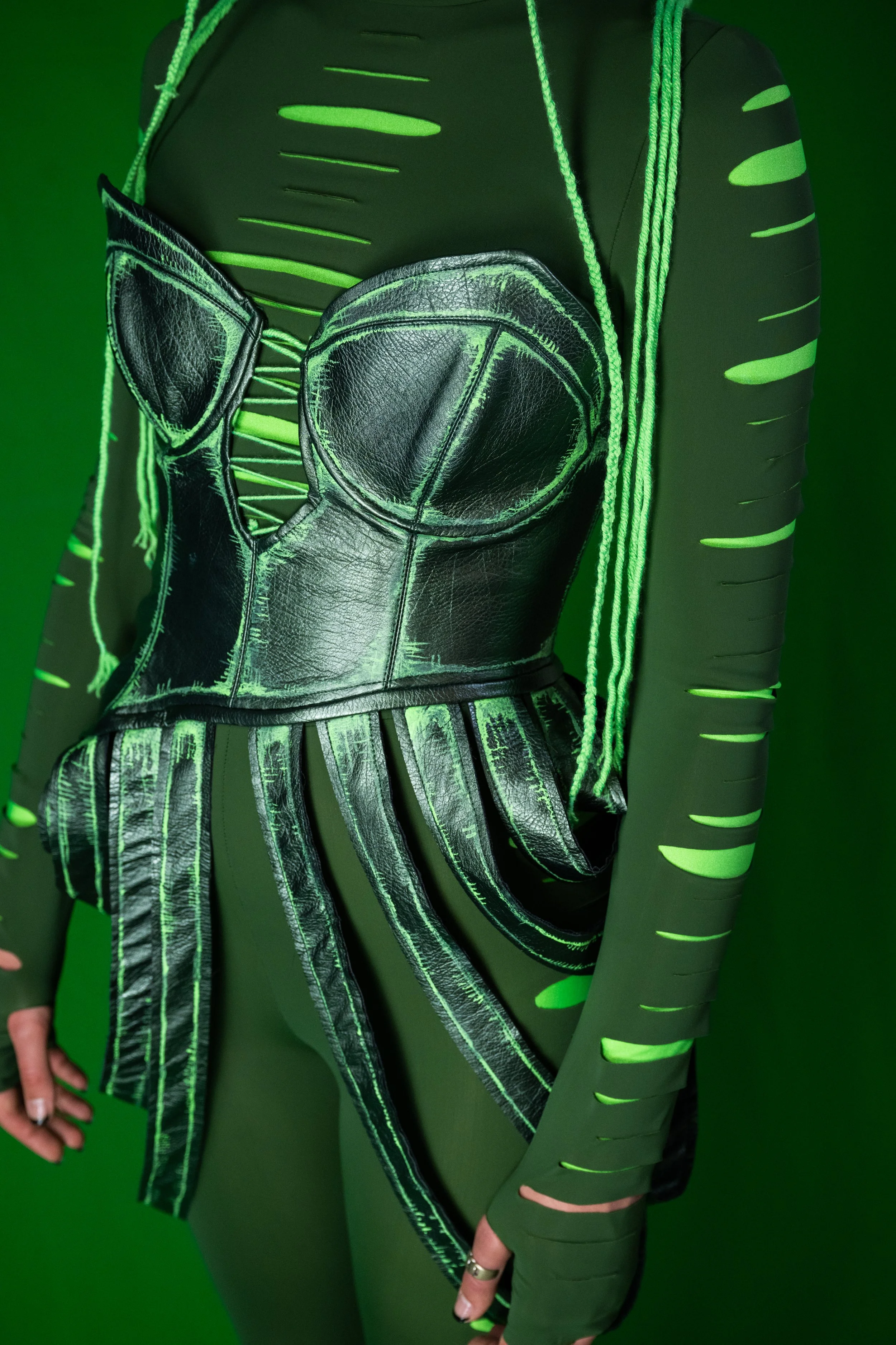 Person wearing a black, leather-like, structured top with neon green accents, and a long sleeve shirt with neon green stripes, standing in front of a green background.