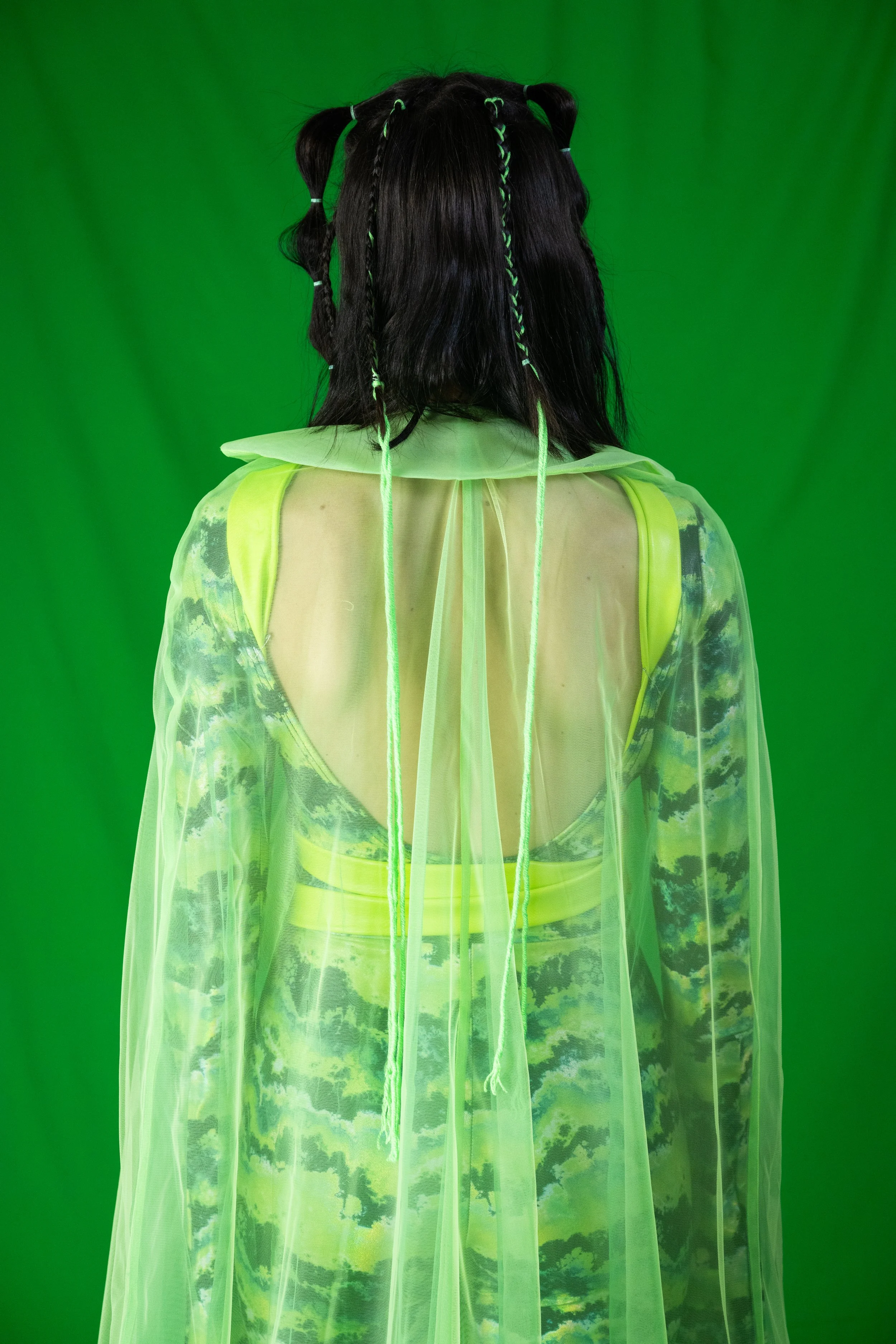 Person with long dark hair styled with green braids, facing away, wearing a sheer green and yellow patterned dress with a bright green neon jacket, standing in front of a green screen.