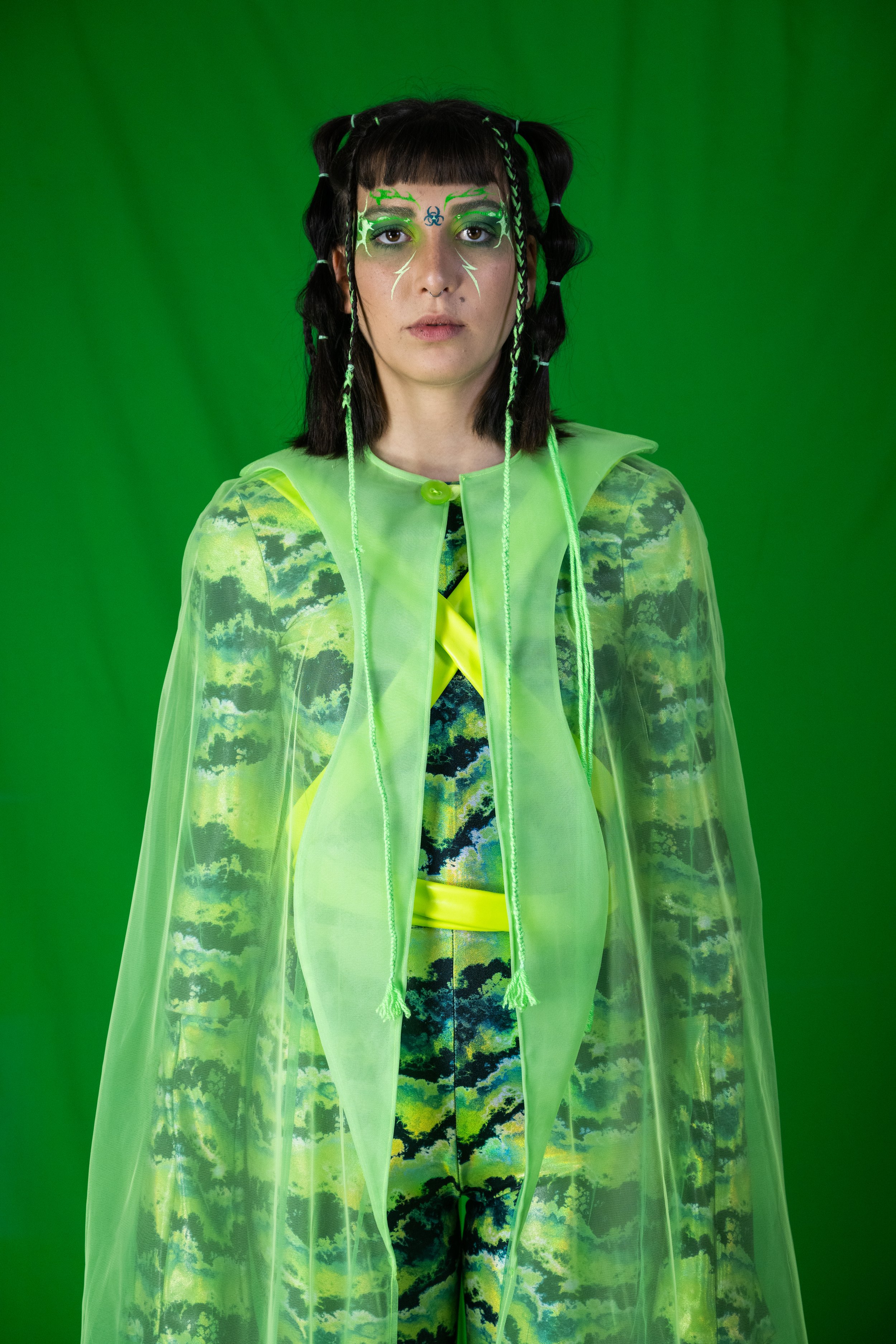 A woman standing in front of a green screen, wearing a neon yellow jacket with shoulder pads, a sheer neon yellow overlay, and rainbow tie-dye clothing underneath. She has black hair styled in pigtails with neon green braids and face paint with neon 