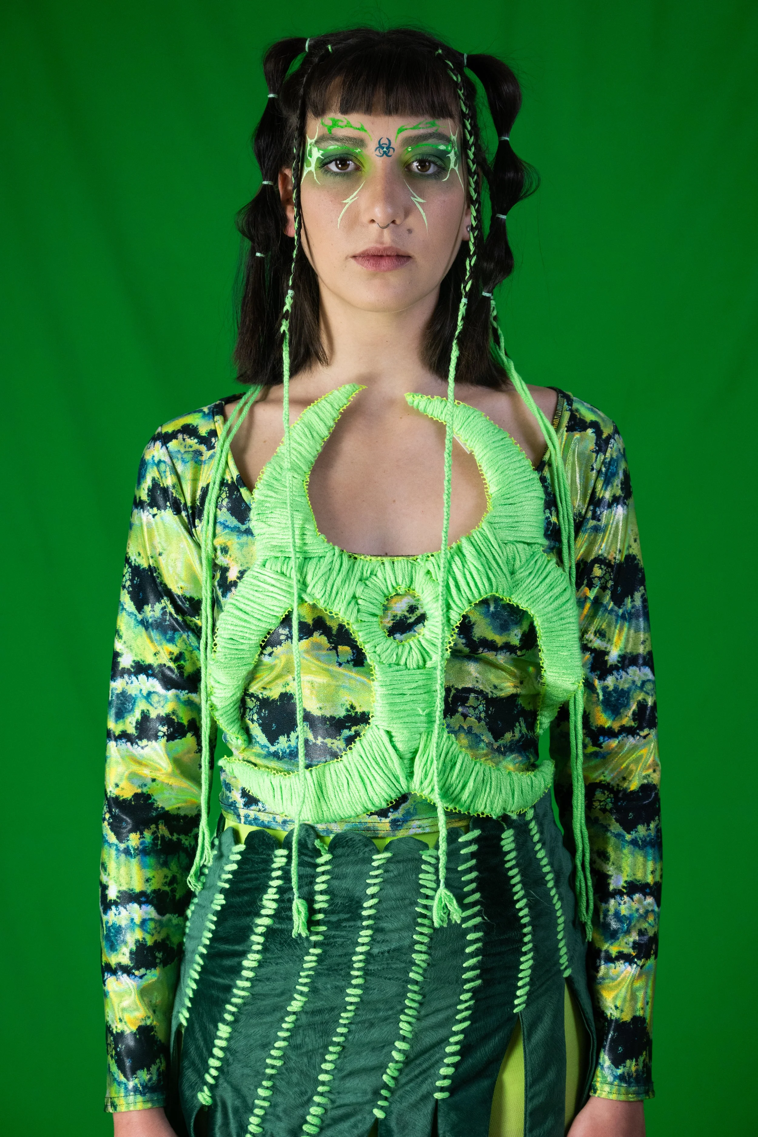 A young woman with dark hair in pigtails styled with green and black hair ties, wearing dramatic green face paint with a biohazard symbol on her forehead, neon green web-like decorative elements on her face, and an outfit blending green and black wit