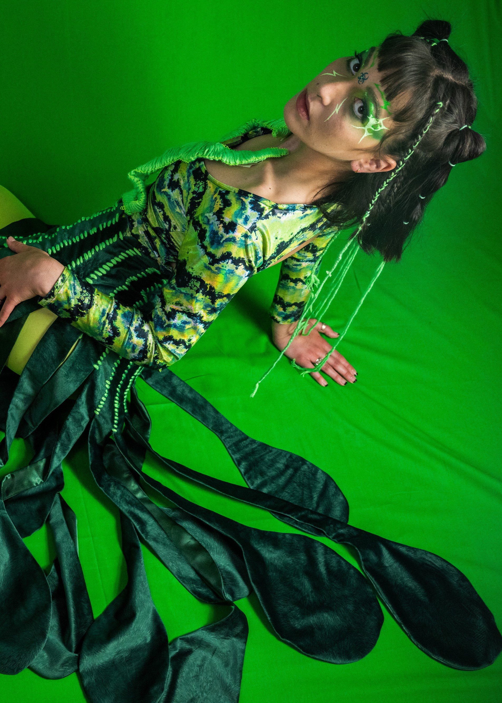 Person with dark hair styled in multiple buns, wearing neon makeup with green and white face paint, a colorful patterned top, and black leather pants, posing against a green screen background.