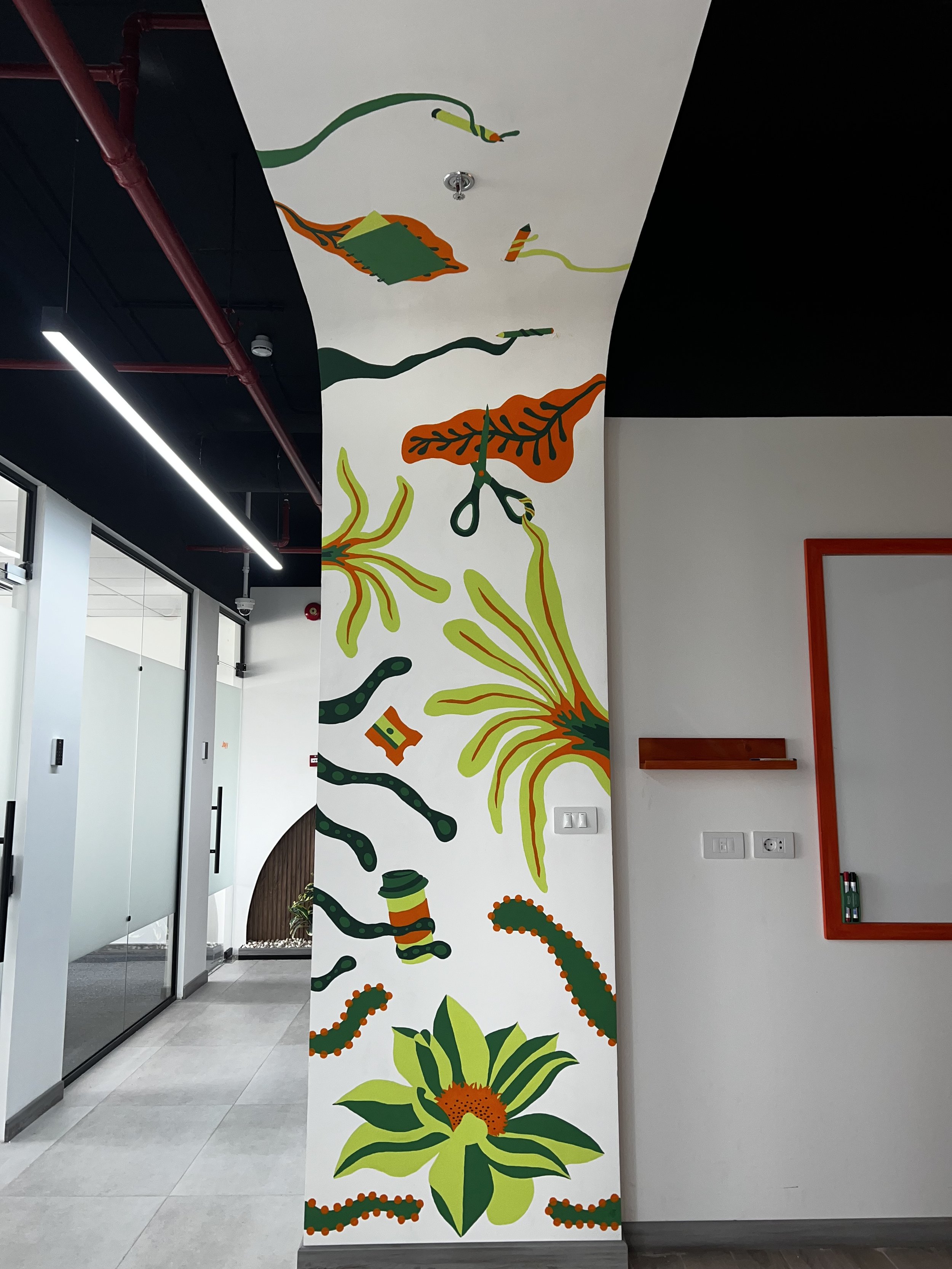 Colorful mural with abstract floral and organic shapes, some resembling leaves, tentacles, and a coffee cup, in green, orange, yellow, and black on a white wall inside a modern office or public building.