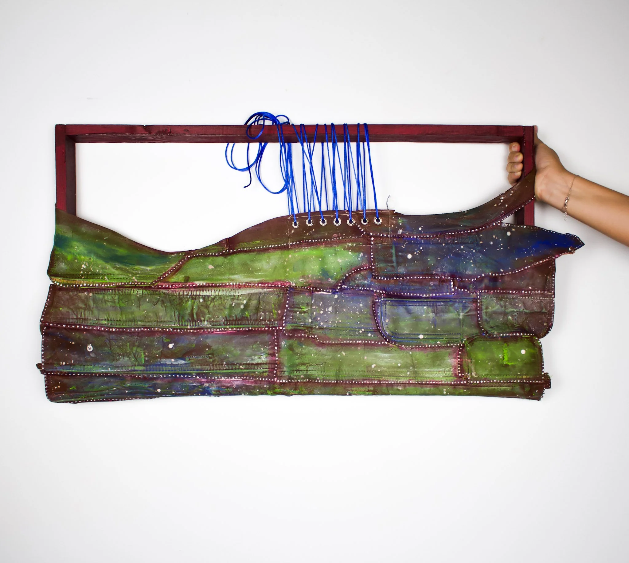 A painting or artistic piece featuring a large whale painted in green and blue colors with white spots, mounted on a red wooden frame, with a person's hand holding the right side of the frame against a plain white wall.