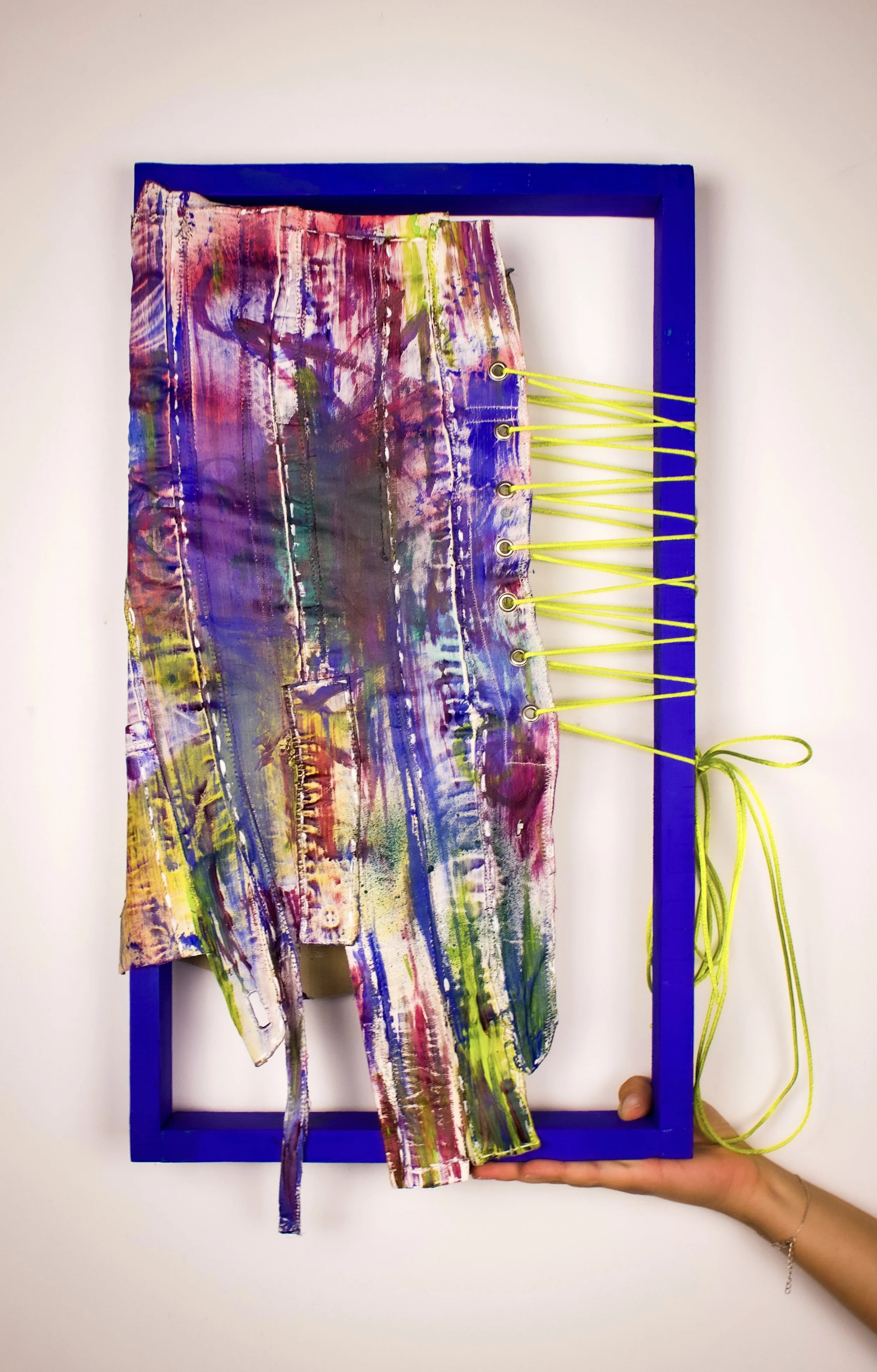 A piece of fabric painted with colorful abstract patterns and tied with yellow strings, held within a blue rectangular frame.