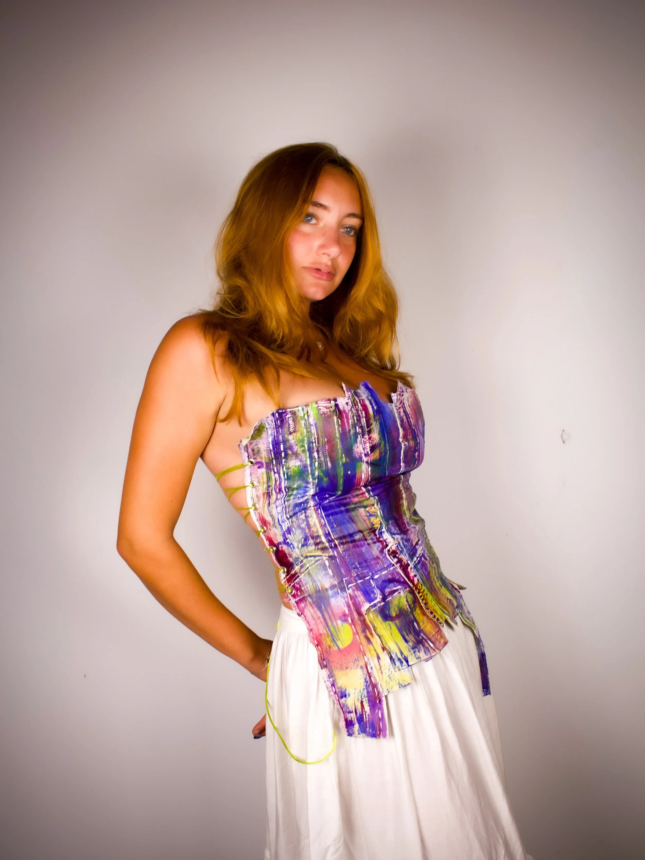 A young woman with red hair and blue eyes wearing a colorful, abstract-patterned top and a flowing white skirt, standing against a plain gray background.