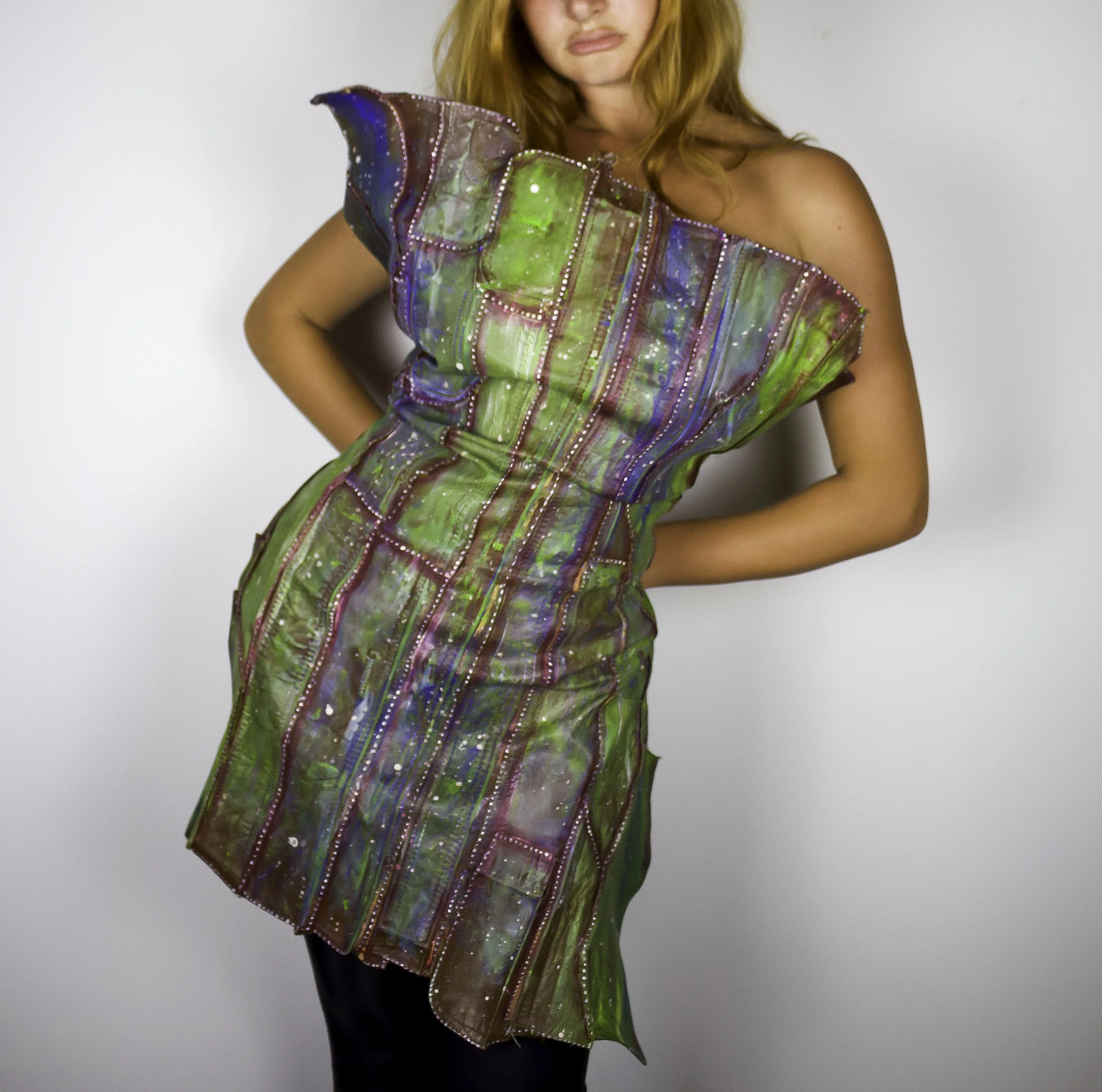 A woman with red hair wearing a colorful, iridescent, strapless dress with vertical stripes and silver embellishments, standing against a plain white background.