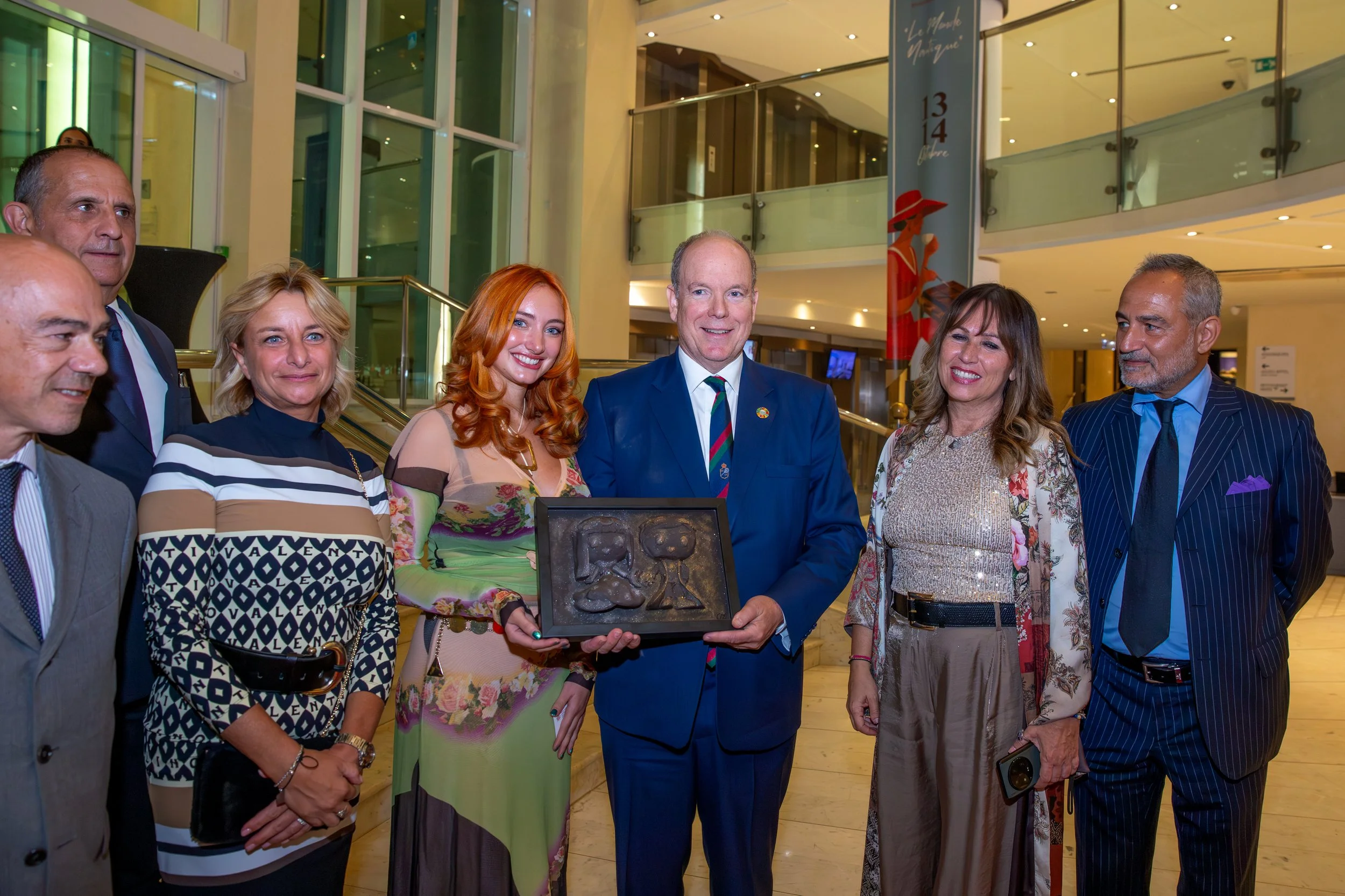 Group of seven people in formal attire standing in an indoor setting, with one person holding a framed object, smiling for a photo.
