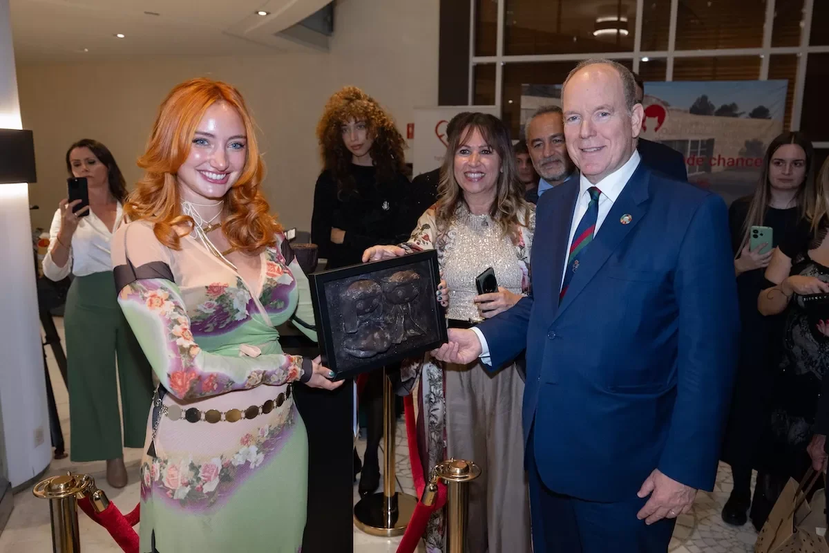 A woman with long red hair wearing a colorful floral dress is holding a framed portrait while a man in a blue suit and striped tie presents it to her. Several people are watching, some taking photos, in an indoor setting.