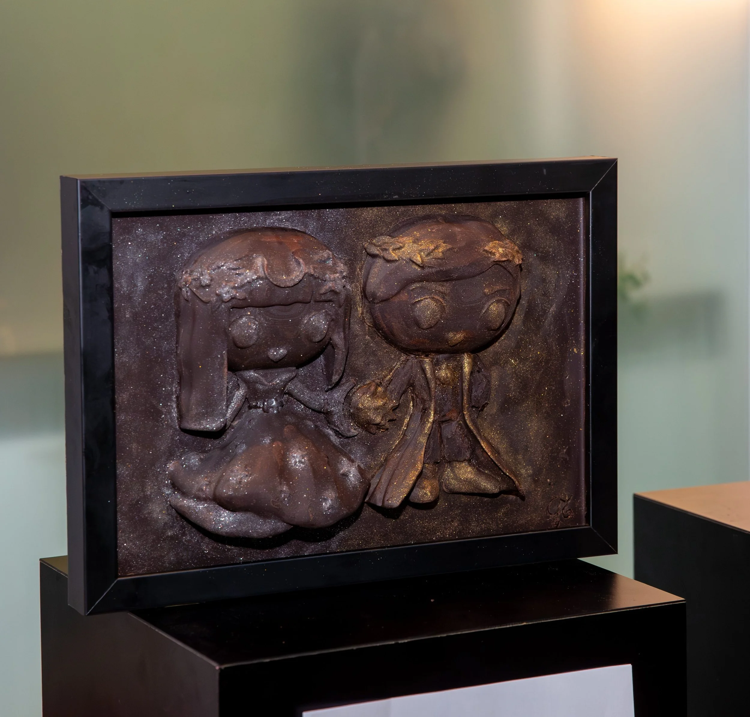 Close-up of a framed artwork featuring detailed sculpted faces with a dark, glittery background, mounted on a black base in an art gallery.