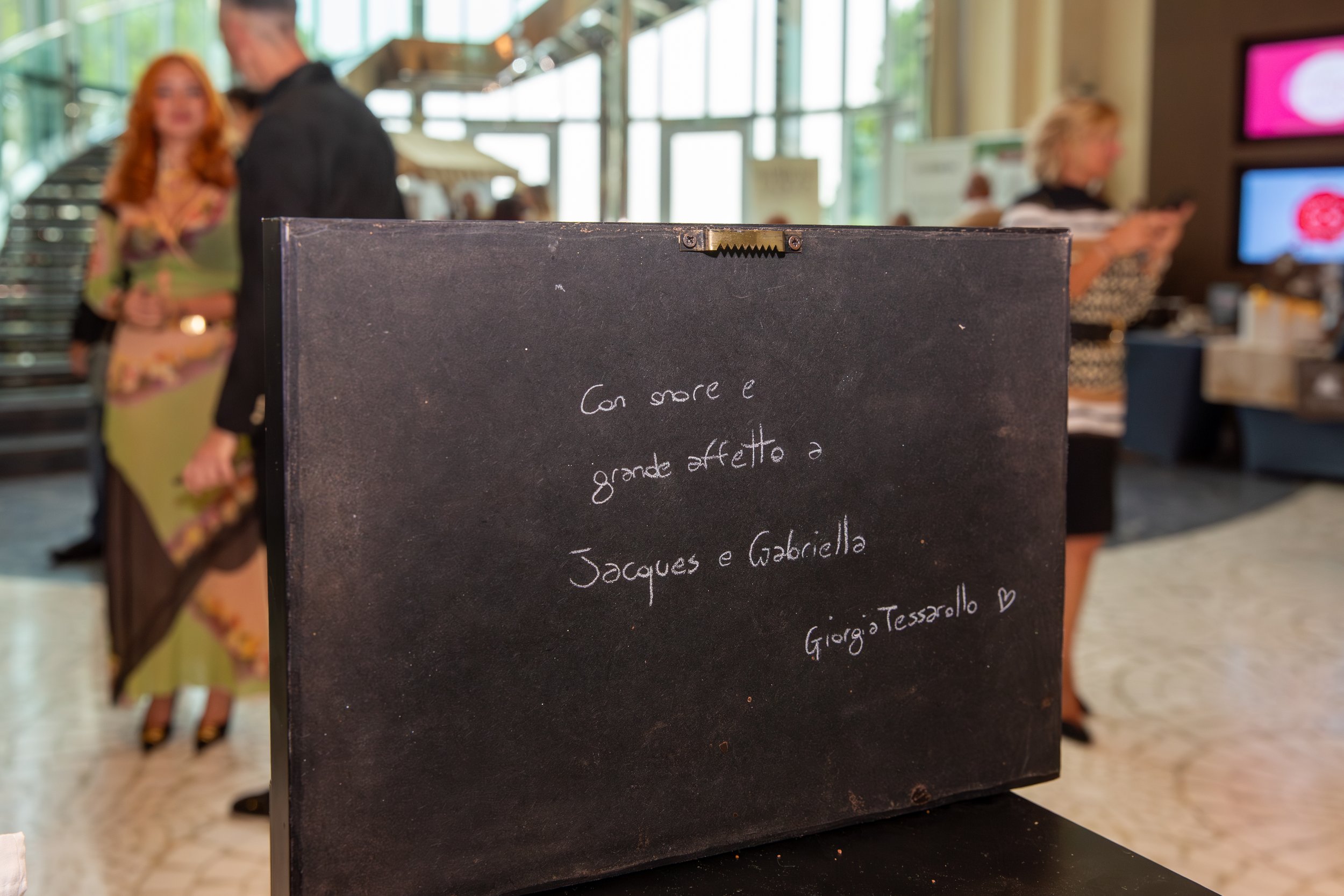A blackboard sign with handwritten Italian names of desserts, placed on a table inside a bright, modern cafe or restaurant. In the background, people are talking and using their phones.