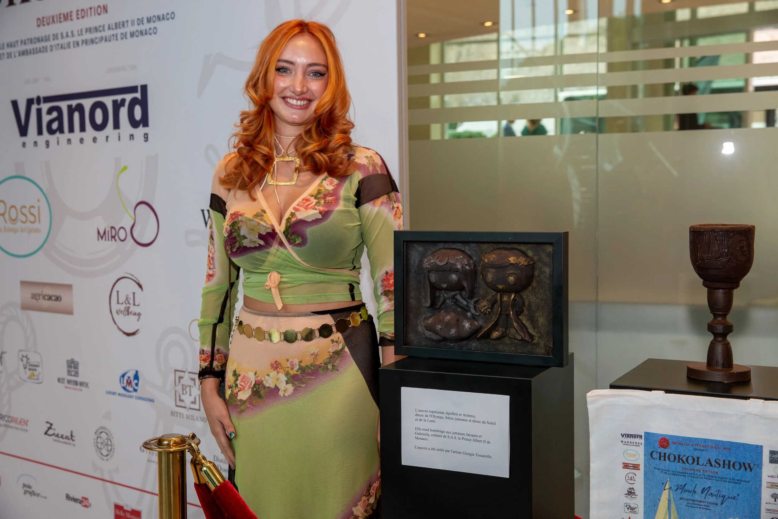A woman with orange hair in a green, floral dress smiling at an art exhibit. Behind her is a poster with logos and text. To her right are sculptures, including a framed artwork of two faces, a wooden sculpture, and a gold-colored object with a red velvet rope barrier.