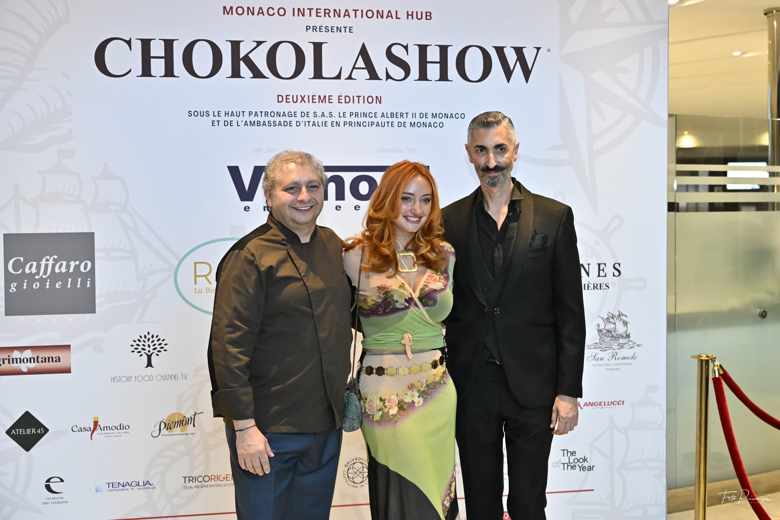 Three people standing in front of a large promotional poster for Chokolashow, a chocolate show event, at Monaco International Hub. The two men are dressed in black suits, and the woman is wearing a colorful, patterned dress. There are various sponsor