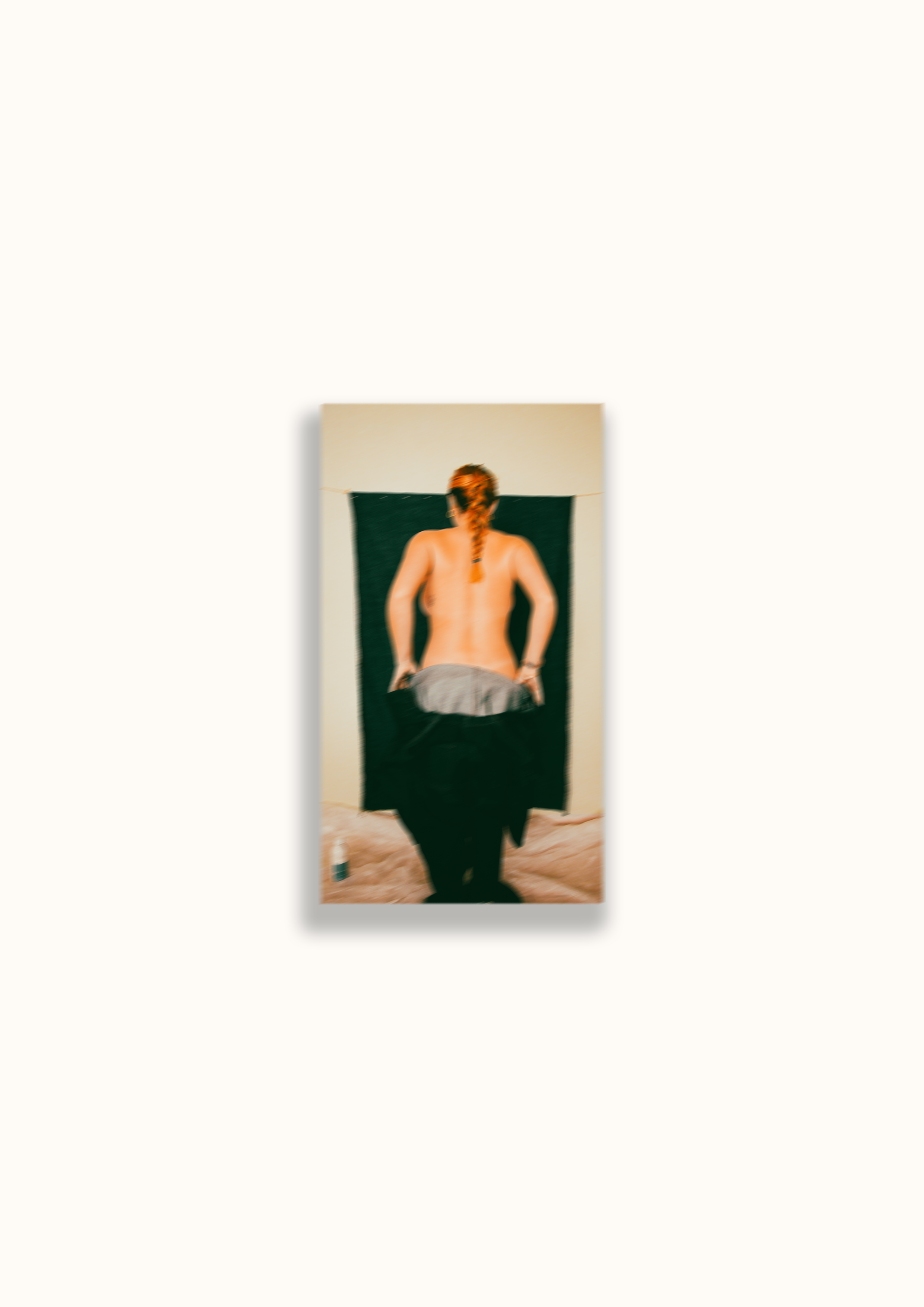 Woman with red hair in braid, topless, standing in front of a black background on a piece of fabric, holding a gray cloth, with her back facing the camera.