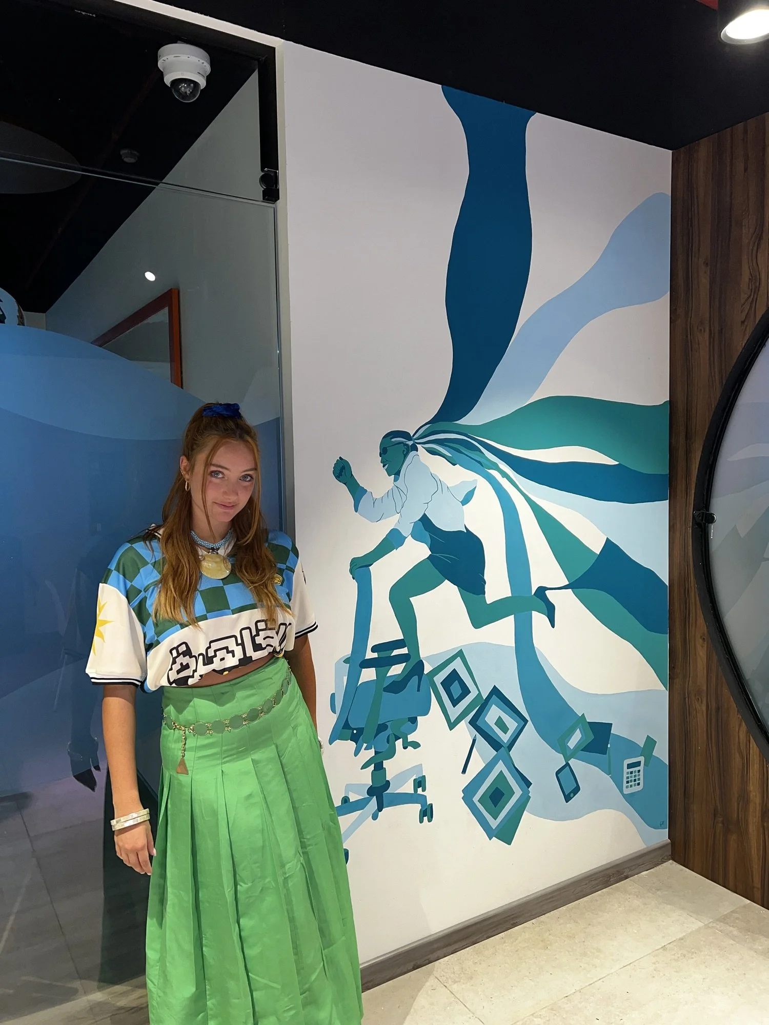 Young woman with long hair, wearing a colorful shirt and a lime green skirt, standing in front of a mural with abstract blue and green designs, in an indoor setting.