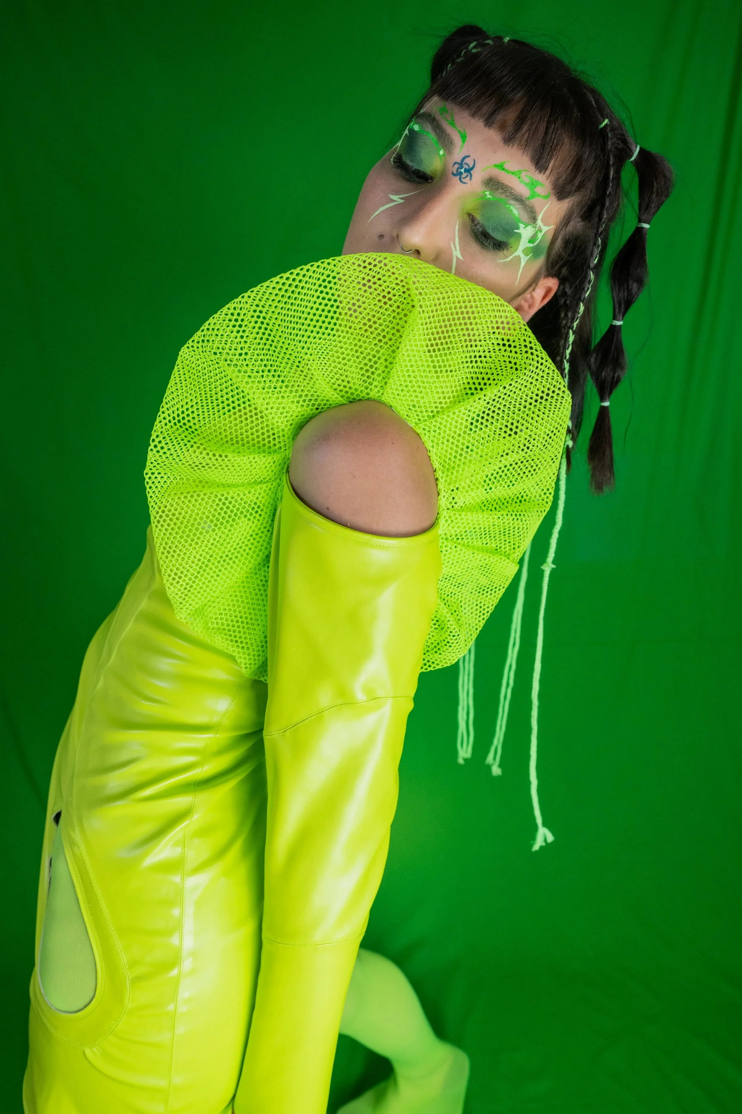 A woman with black hair styled in pigtails with white hair ties, vibrant green and blue face makeup, wearing a neon green mesh top with cutouts on the shoulder, and neon green shiny pants, standing against a green screen background.