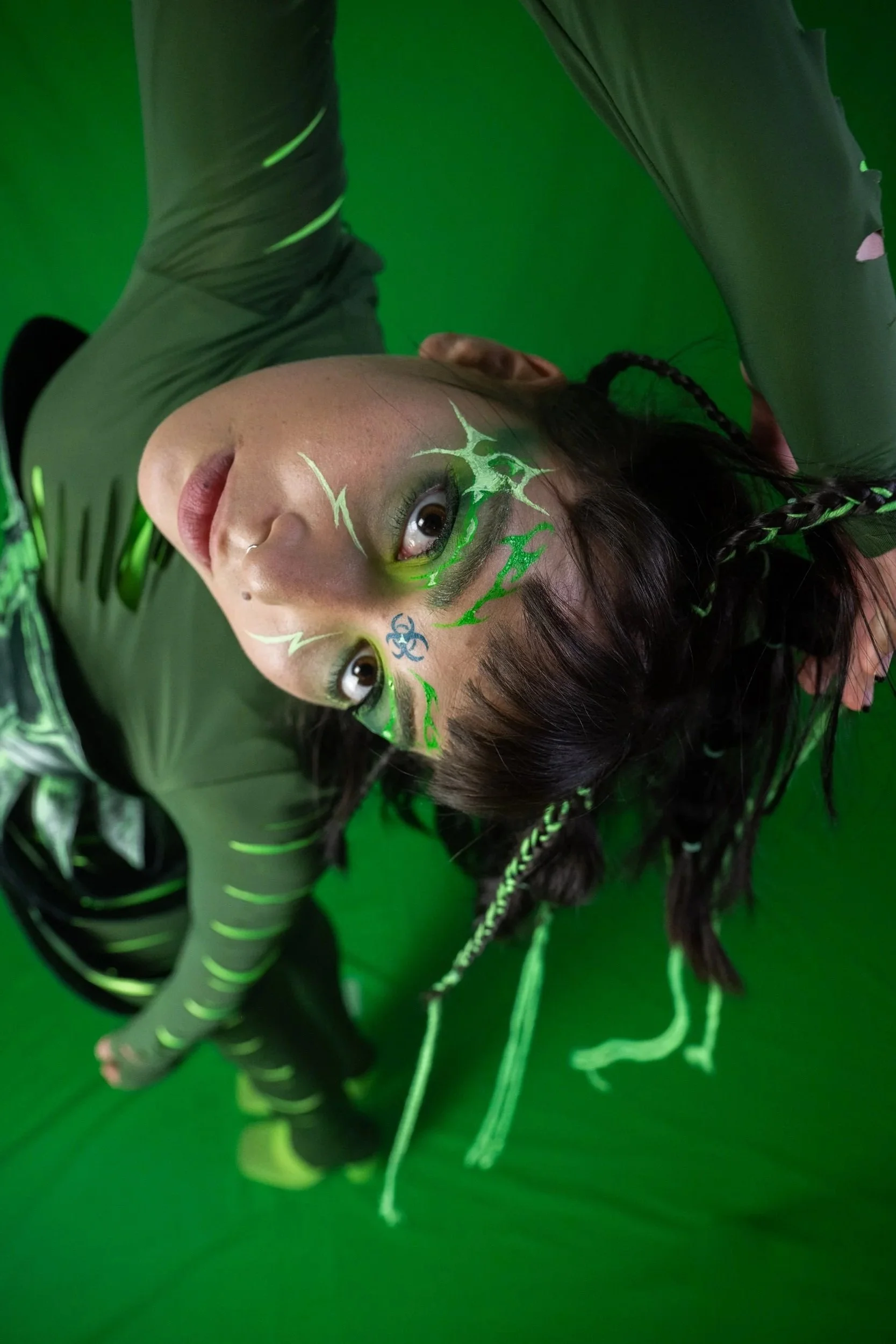 A woman with face paint and accessories posed in a green screen studio. She is wearing a black outfit with neon green accents, with long black braided hair.