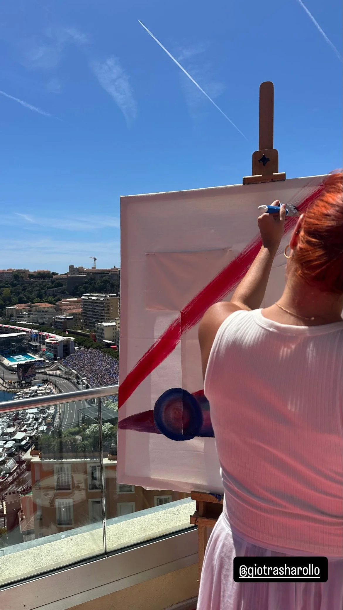A woman with red hair painting on a canvas outdoors with a city view in the background.