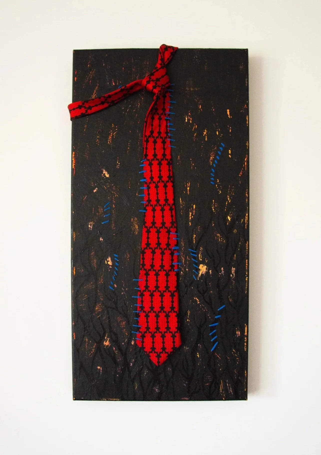 A mixed media art piece featuring a black textured background with a red and black patterned fabric piece tied in a knot at the top, modified with blue stitching along the edges, mounted vertically on a white wall.