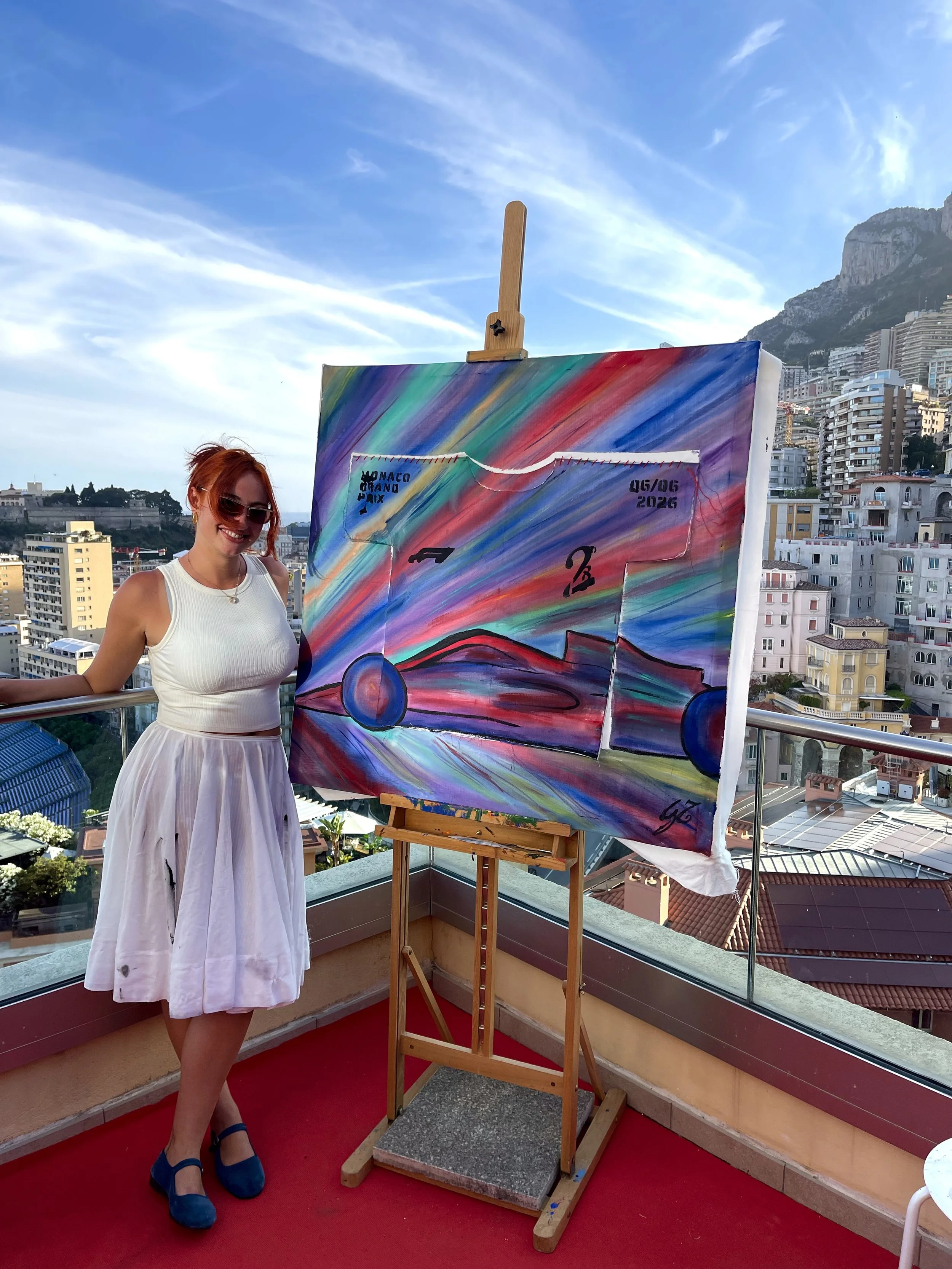 A woman with red hair and sunglasses smiling at an outdoor art exhibition, standing next to a colorful abstract painting on an easel on a rooftop with city buildings and mountains in the background.