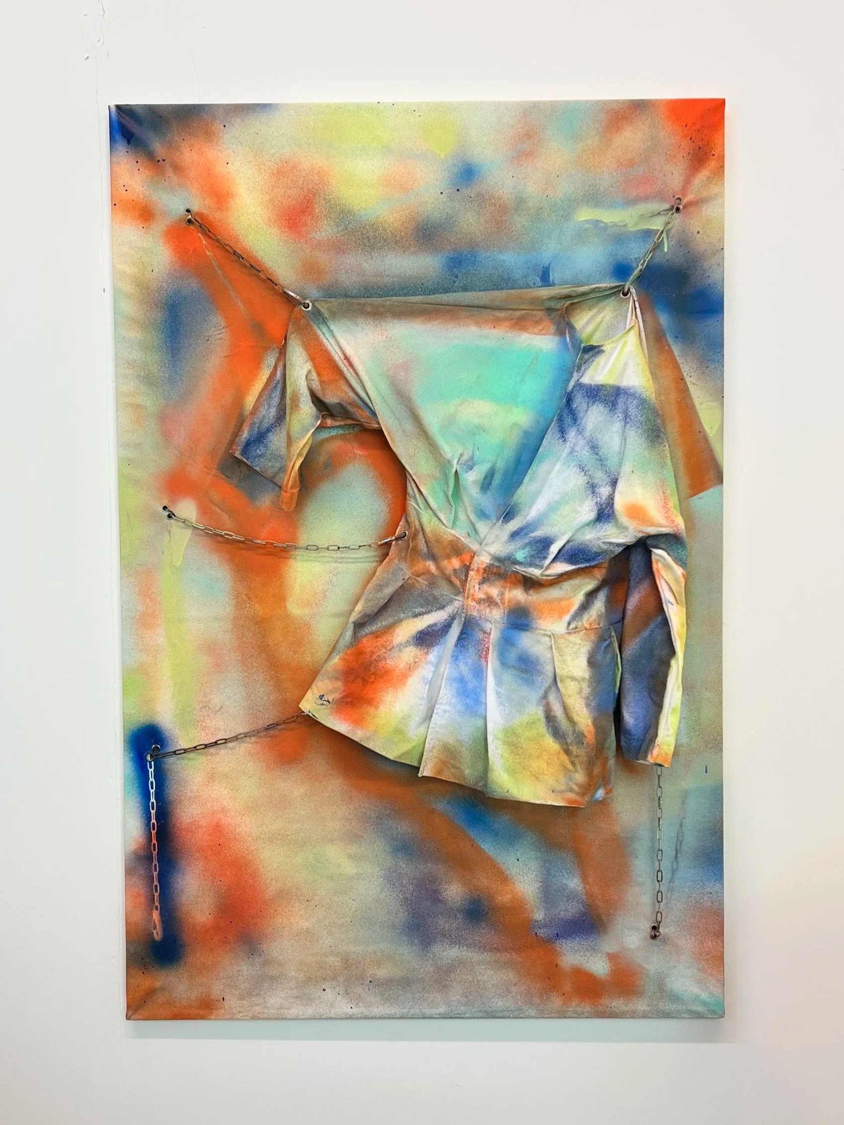 Mixed media artwork with spray paint background in vibrant colors and fabric or paper elements attached with chains, creating a textured, layered composition.