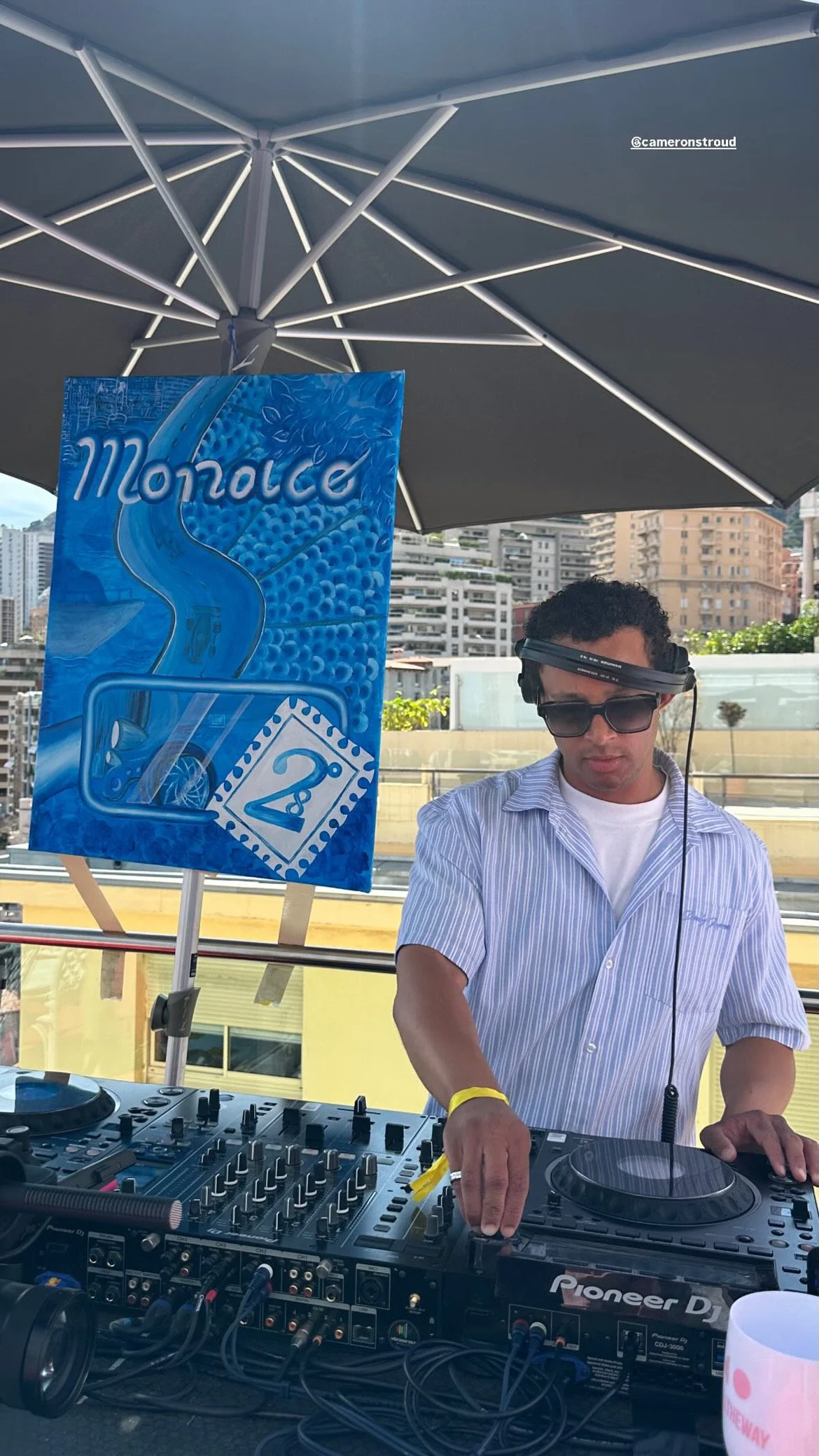 A DJ wearing headphones and sunglasses performs at an outdoor event, using DJ equipment with city buildings in the background.