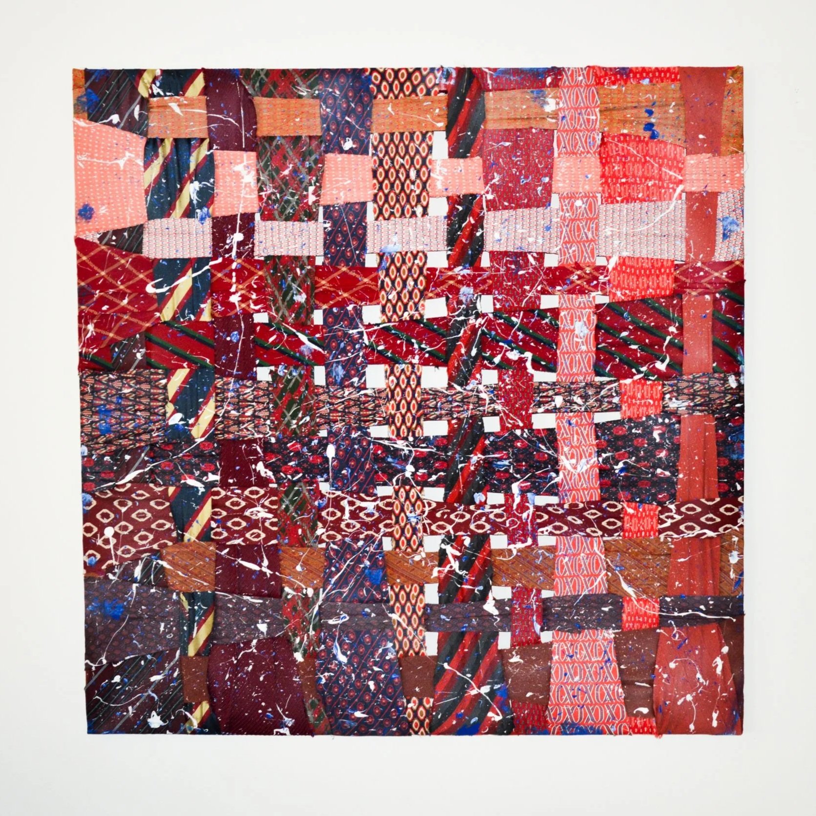 Colorful woven patchwork art made from various fabric strips in red, blue, black, and orange, with paint splatters and splashes across the surface, mounted on a white background.
