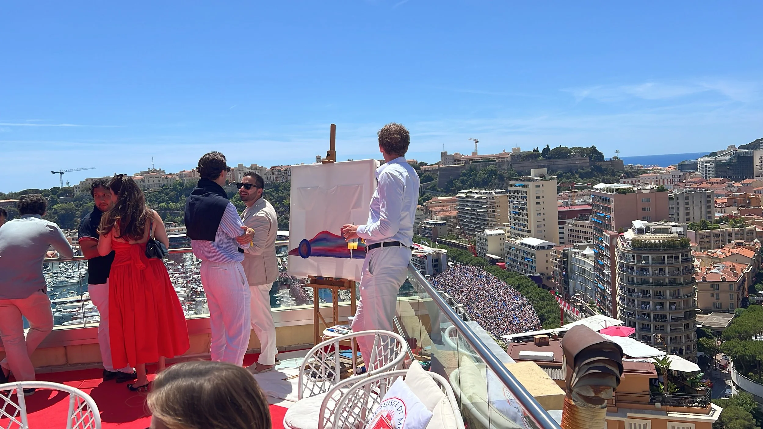 People gathered on a rooftop terrace overlooking a city with a hillside fortress, some of them engaged in conversation, with painting supplies nearby, under a clear blue sky.